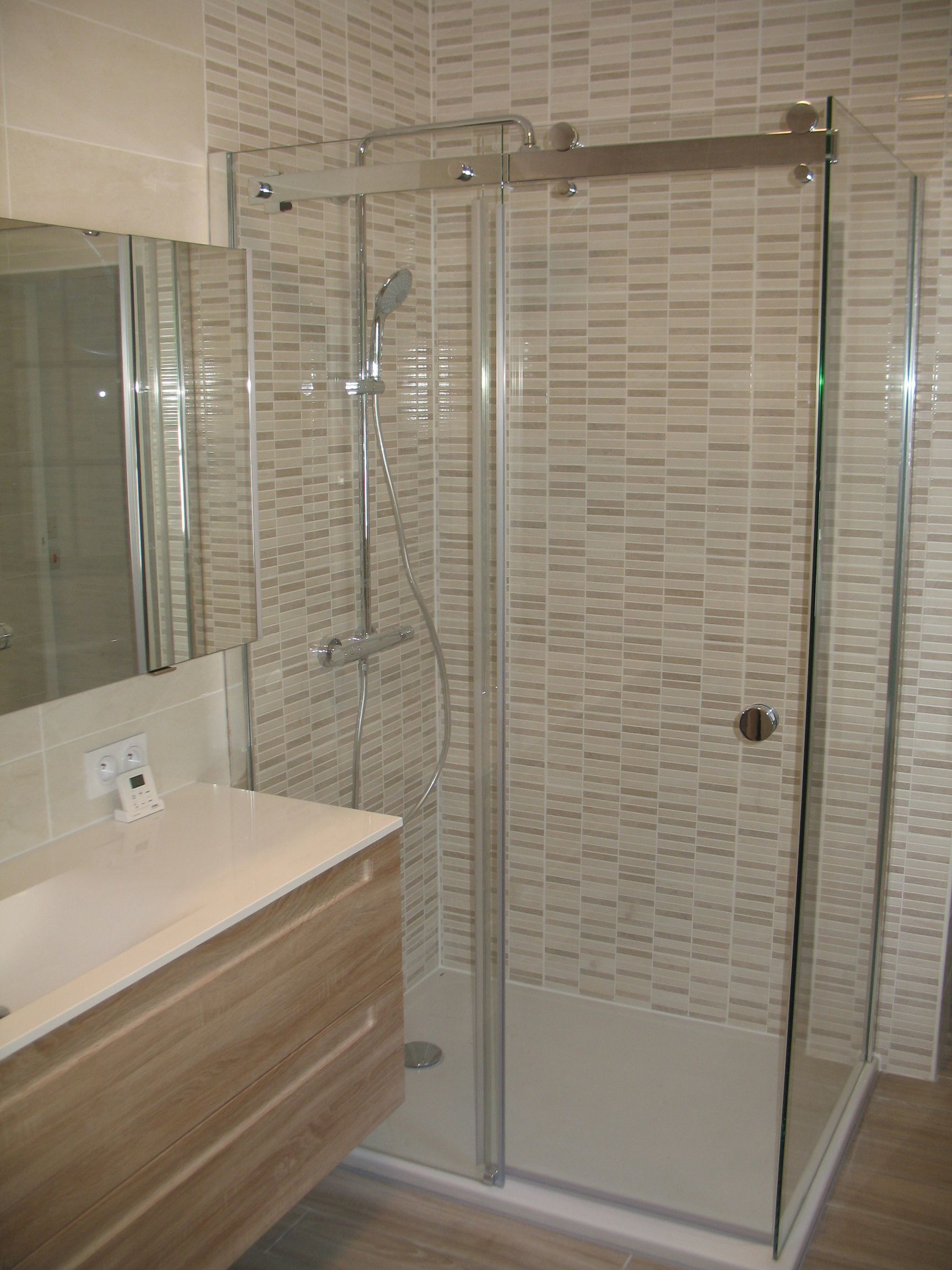 Cabine de douche avec portes en verre, luminaires argentés, murs en carrelage de couleur claire et vanité en bois.