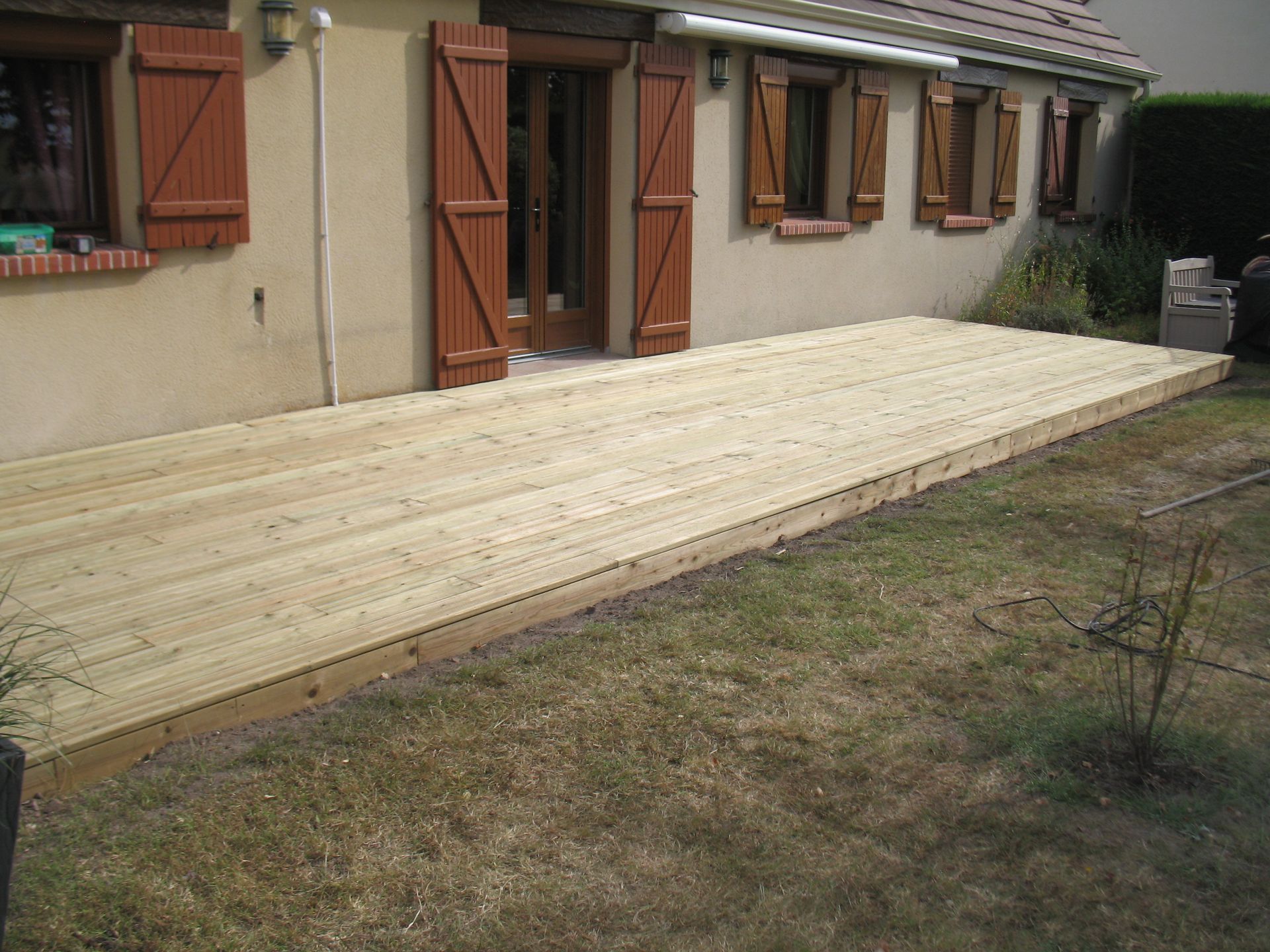 Terrasse en bois prolongeant une maison beige aux volets marron ; construite sur une pelouse herbeuse.
