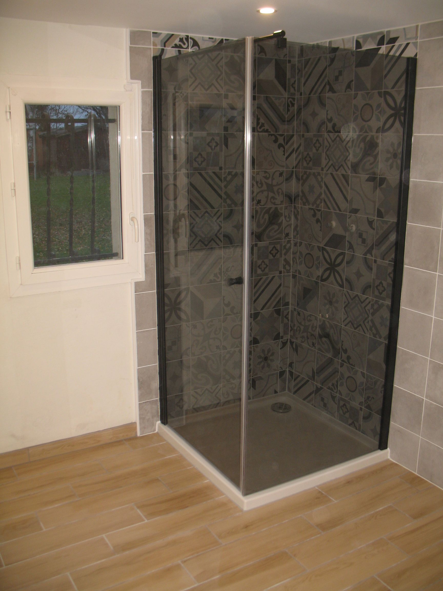 Douche d'angle avec carrelage gris foncé à motifs, base grise, cadre noir et fenêtre.