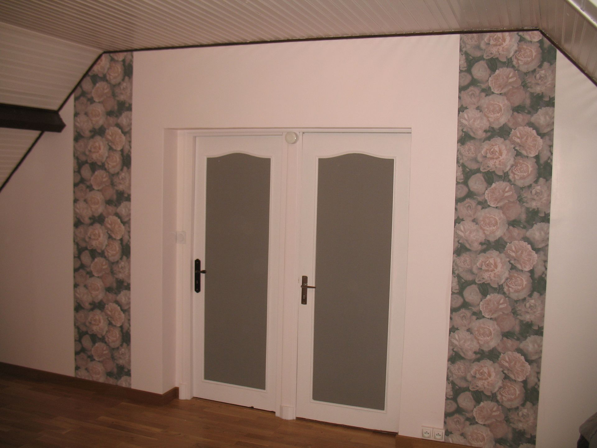 Portes doubles blanches avec verre dépoli, encadrées par des colonnes en papier peint floral, dans une pièce avec parquet.