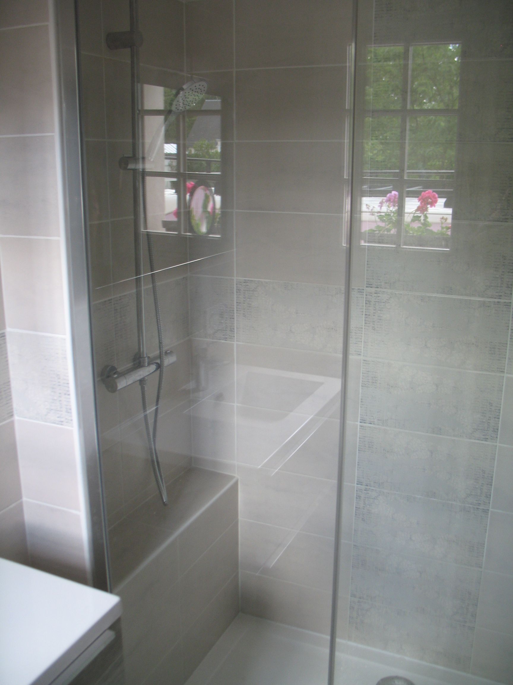 Cabine de douche avec porte vitrée, carrelage gris, pommeau de douche et fenêtre avec fleurs.