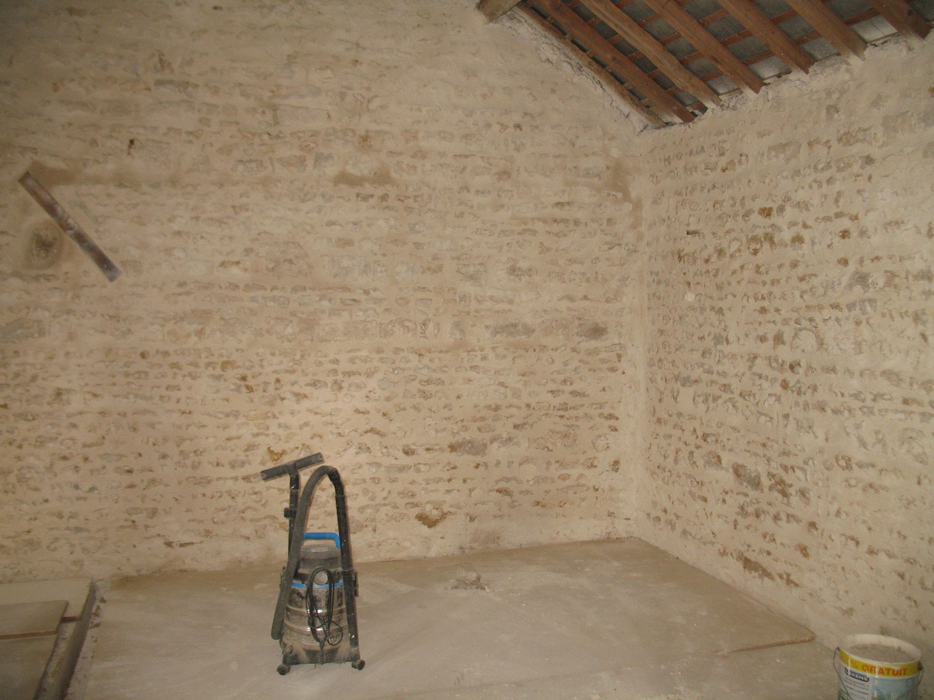 Vue intérieure d'une pièce aux murs de pierre, avec une échelle et des poutres apparentes au plafond.