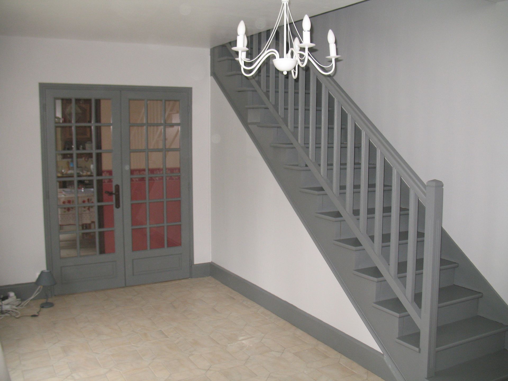 Escalier et porte gris dans une pièce ornée d'un lustre. Les murs sont blancs et les boiseries grises.