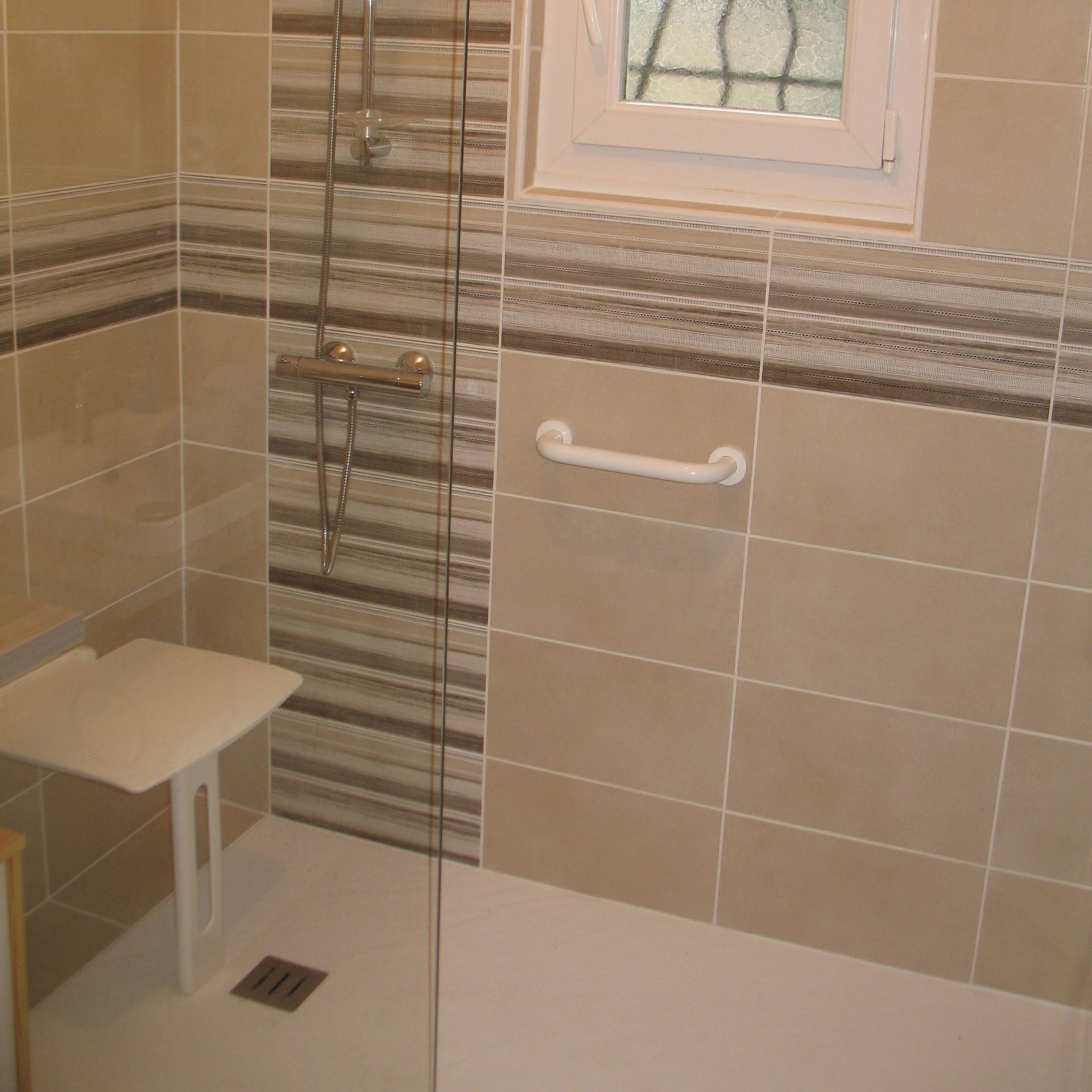 Cabine de douche accessible avec barre d'appui, siège rabattable et fenêtre. Carrelage beige à rayures claires et foncées.
