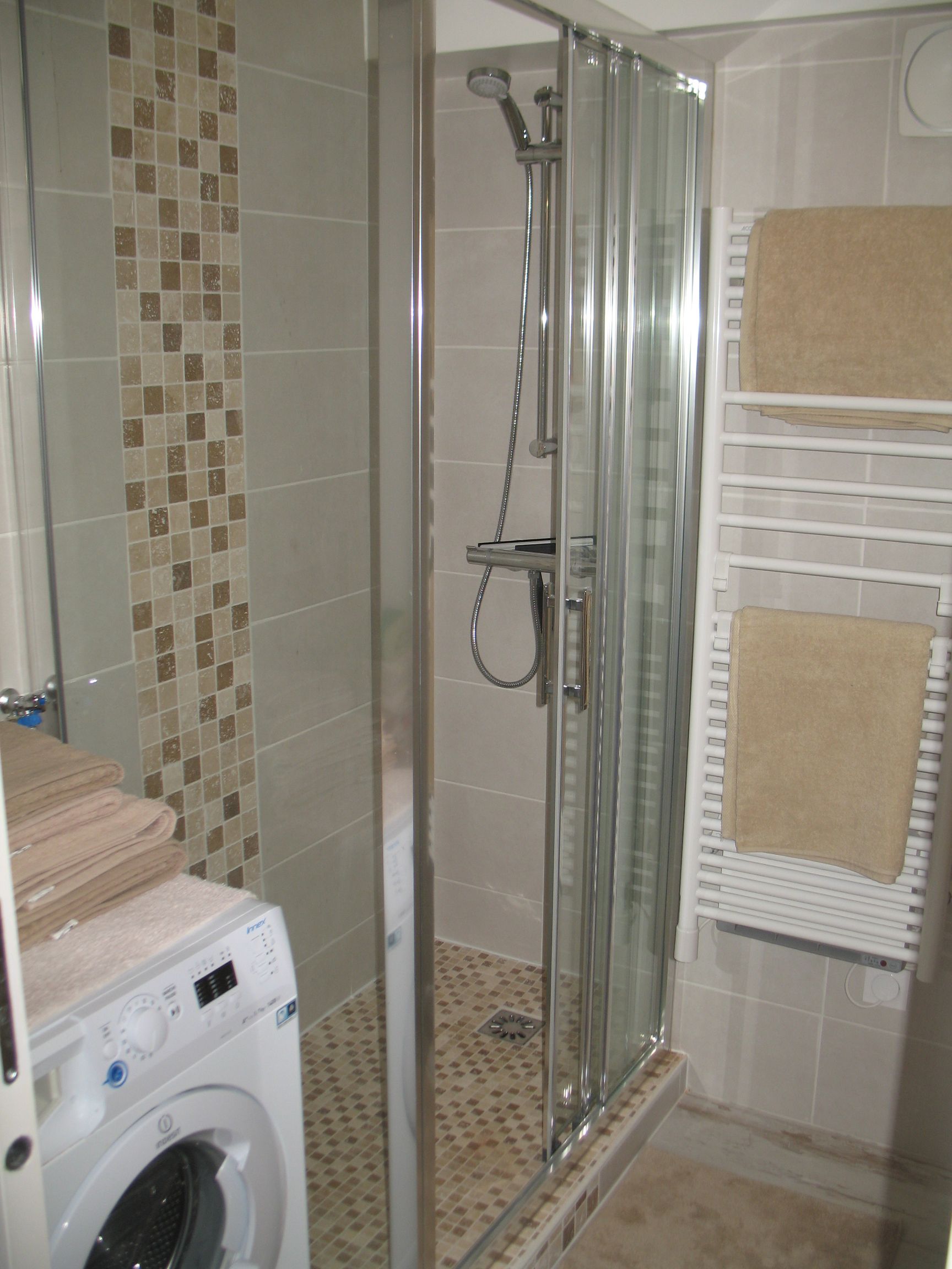 Petite salle de bains avec douche, lave-linge et sèche-serviettes. Couleurs beige et gris.