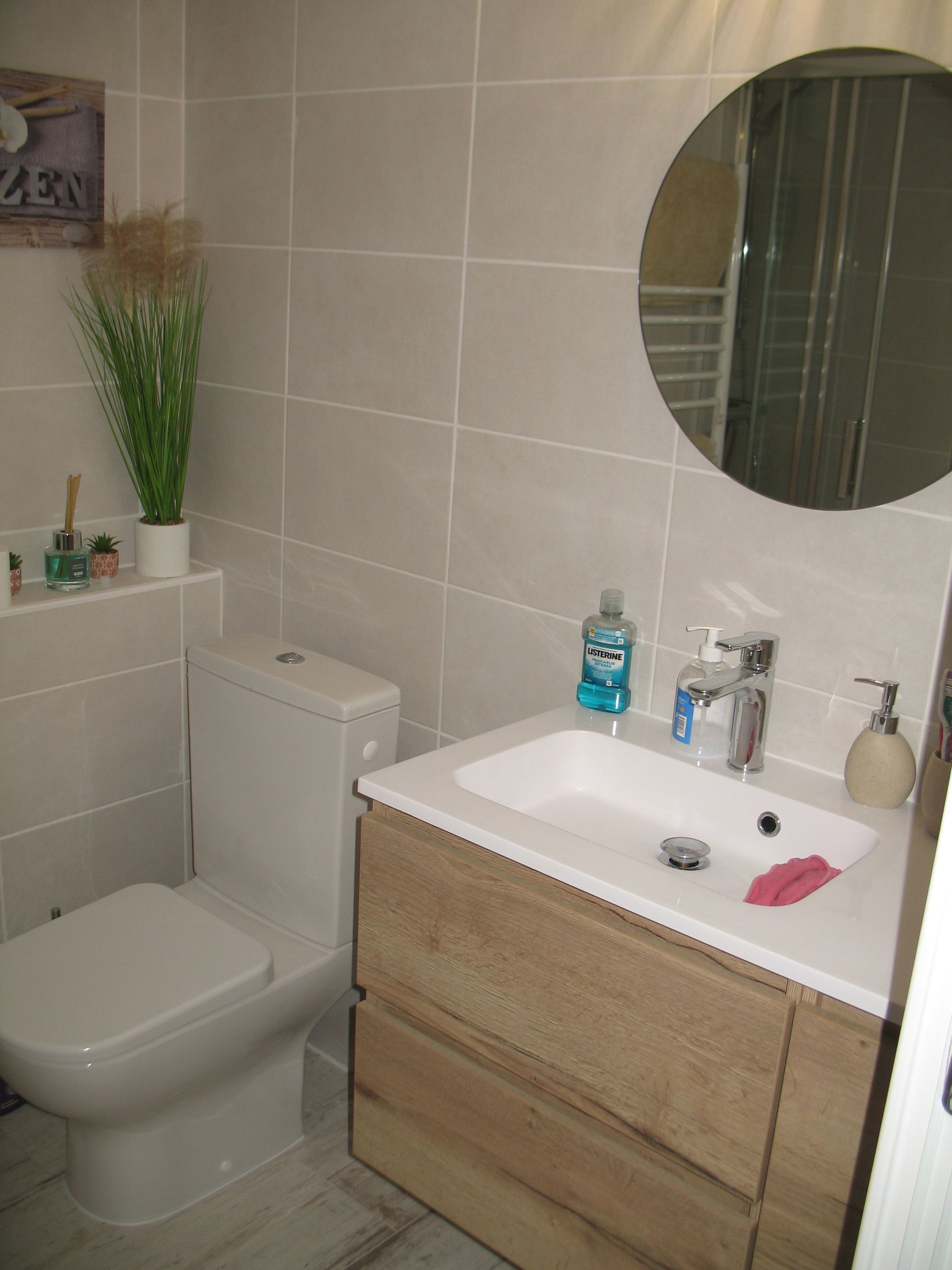 Salle de bain avec murs carrelés gris clair, toilettes, lavabo et meuble-lavabo en bois. Un miroir rond est suspendu.