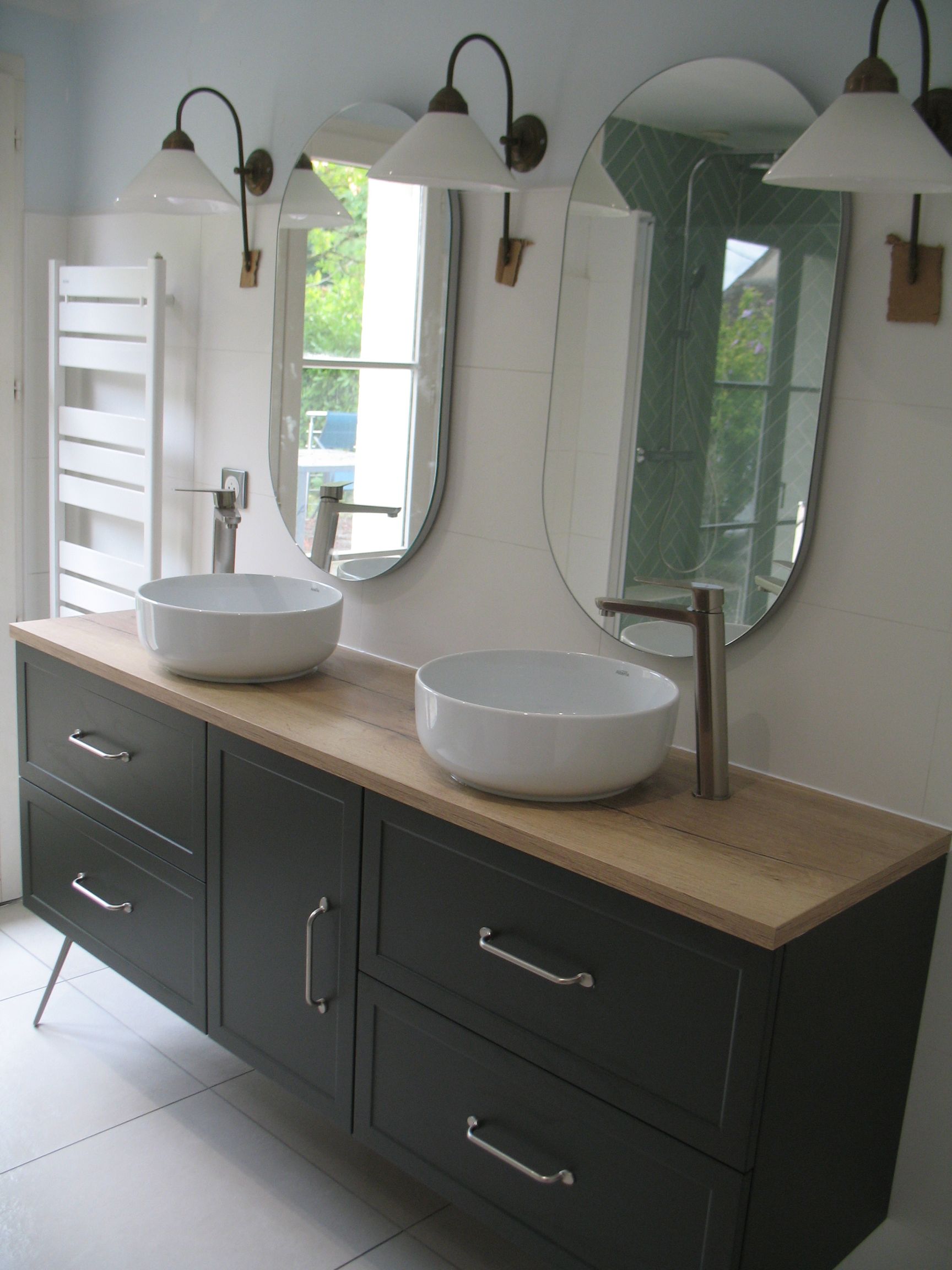 Salle de bain avec double vasque; armoire grise, comptoir en bois, miroirs ovales, lavabos et appliques murales.