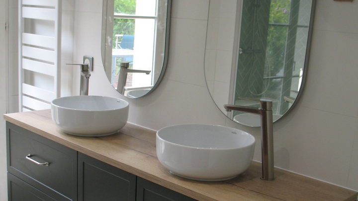 Meuble à lavabo avec deux vasques blanches, miroirs ovales et plan de travail en bois clair. Meuble bas foncé.