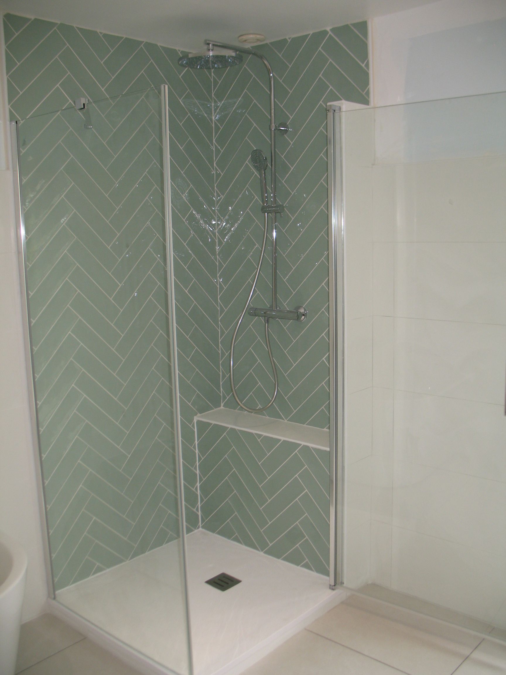 Douche avec carrelage à chevrons vert clair, porte en verre et base et murs de douche blancs.