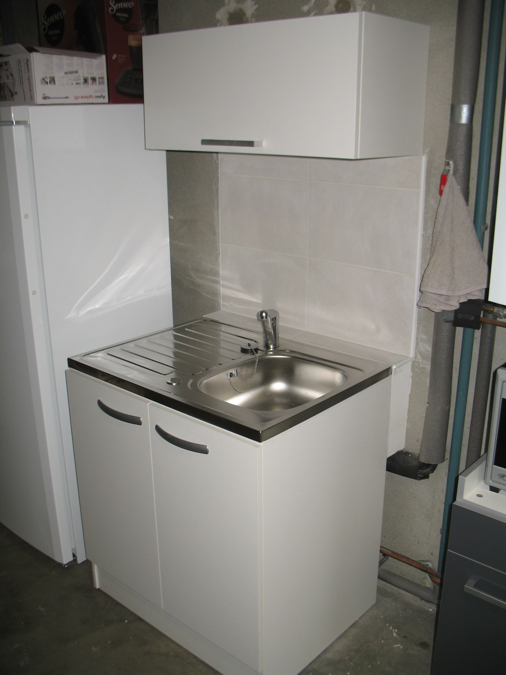 Meuble utilitaire blanc avec rangement supérieur, robinet et réfrigérateur, placé contre un mur neutre.