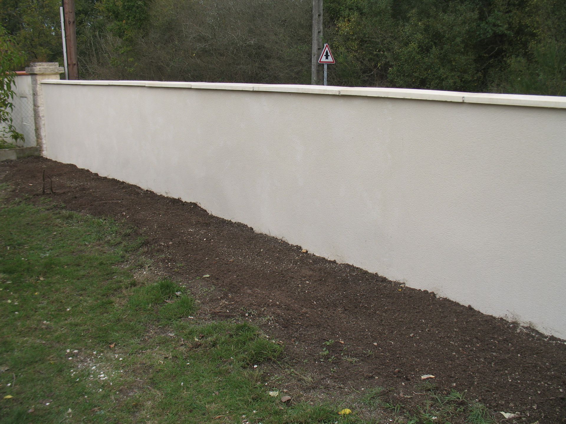 Un long mur de couleur claire borde une bande de terre sombre et d'herbe.