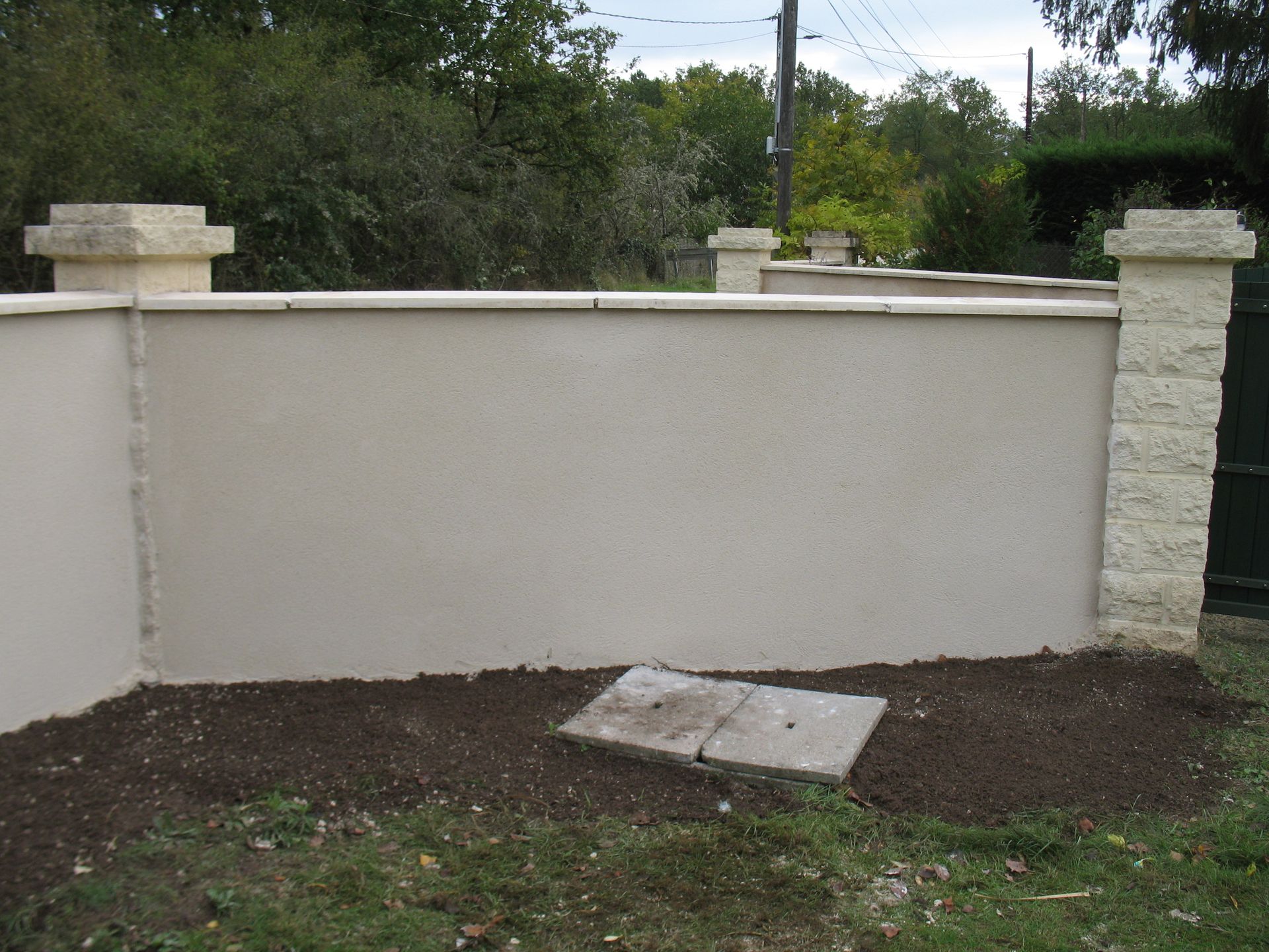 Un mur en stuc beige avec des piliers en pierre. Une petite couverture en béton repose au sol devant le mur.