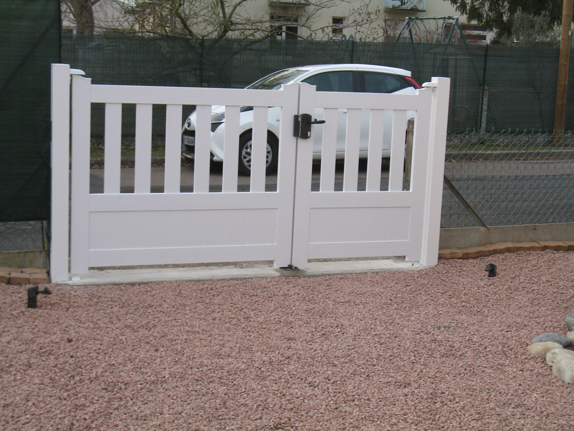 Portail double blanc, avec une voiture blanche en arrière-plan, posé sur un lit de gravier rose.