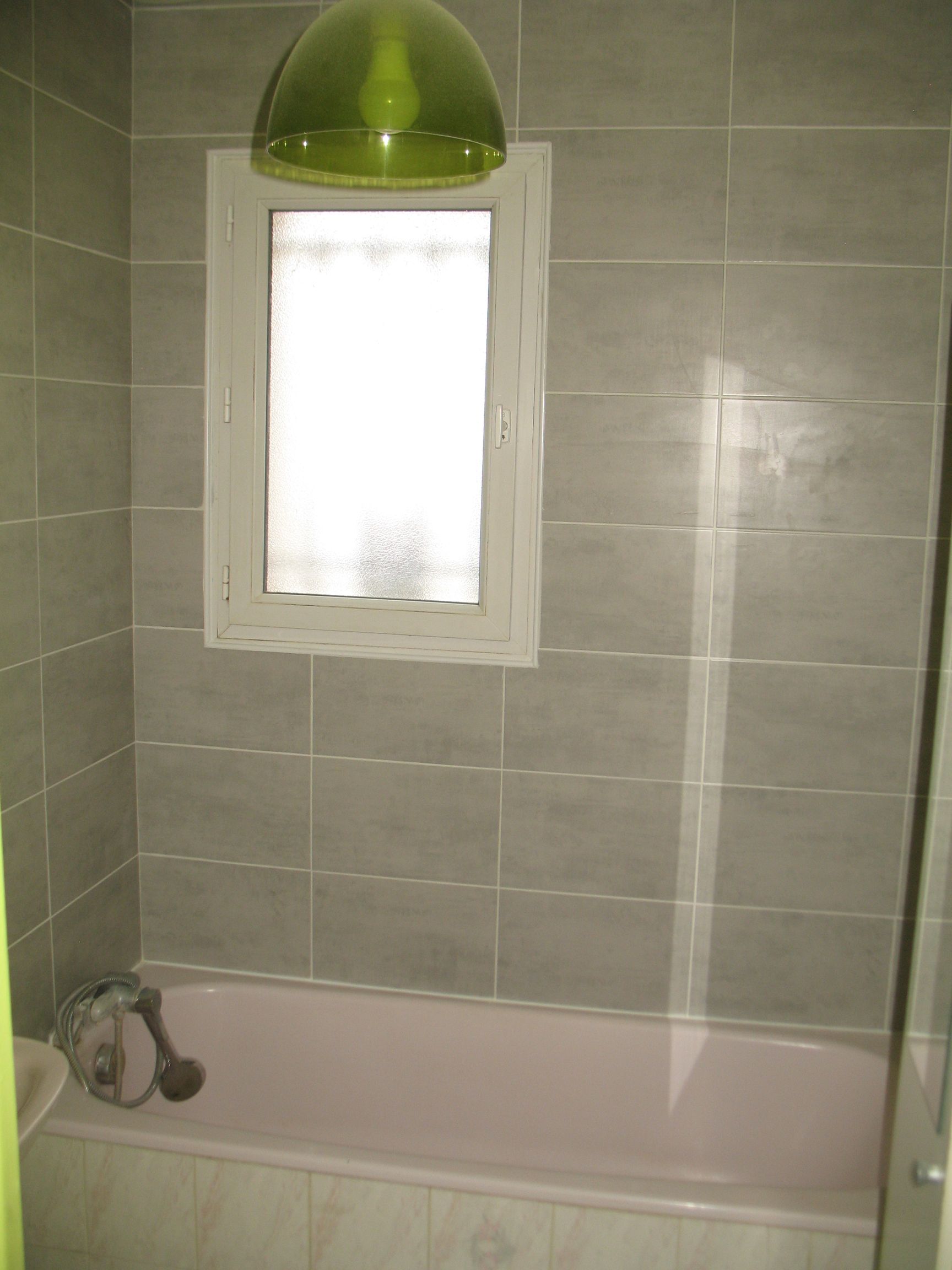 Salle de bain avec des murs carrelés gris, une baignoire rose et une fenêtre avec un luminaire vert au-dessus.