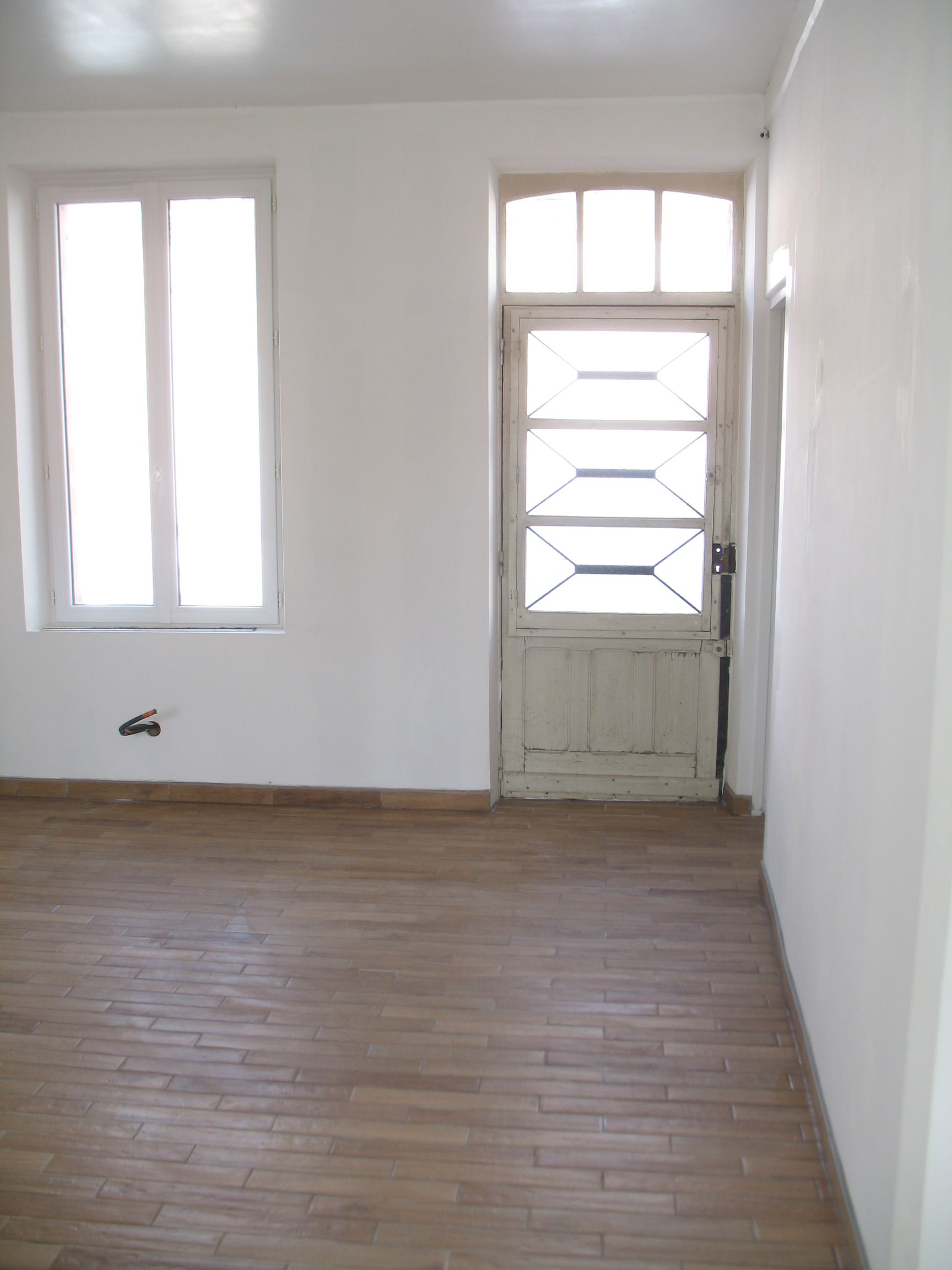 Pièce vide avec fenêtre et porte, parquet, murs blancs et lumière naturelle.