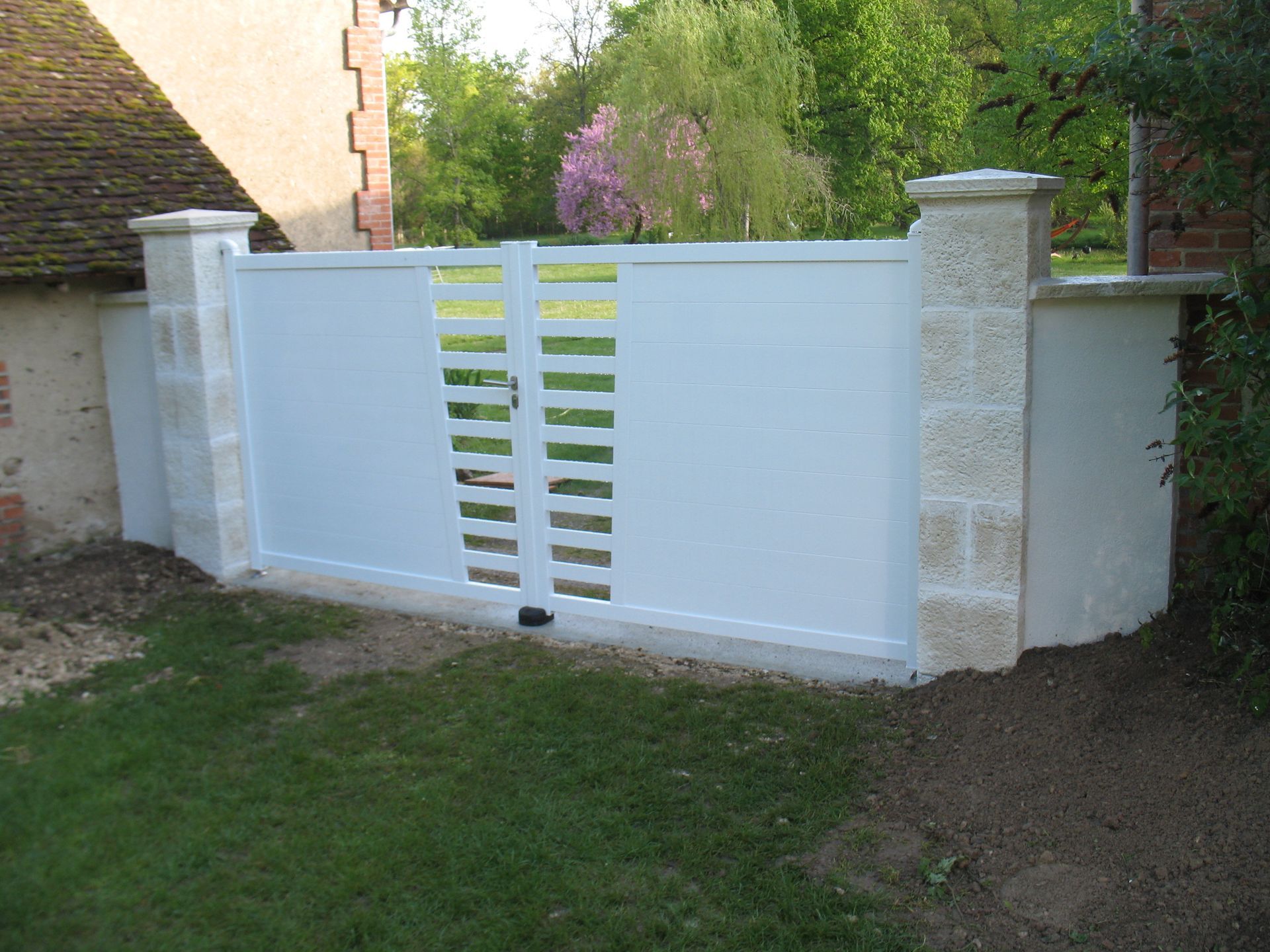 Portail blanc avec découpes horizontales entre piliers en pierre, dans une cour herbeuse.