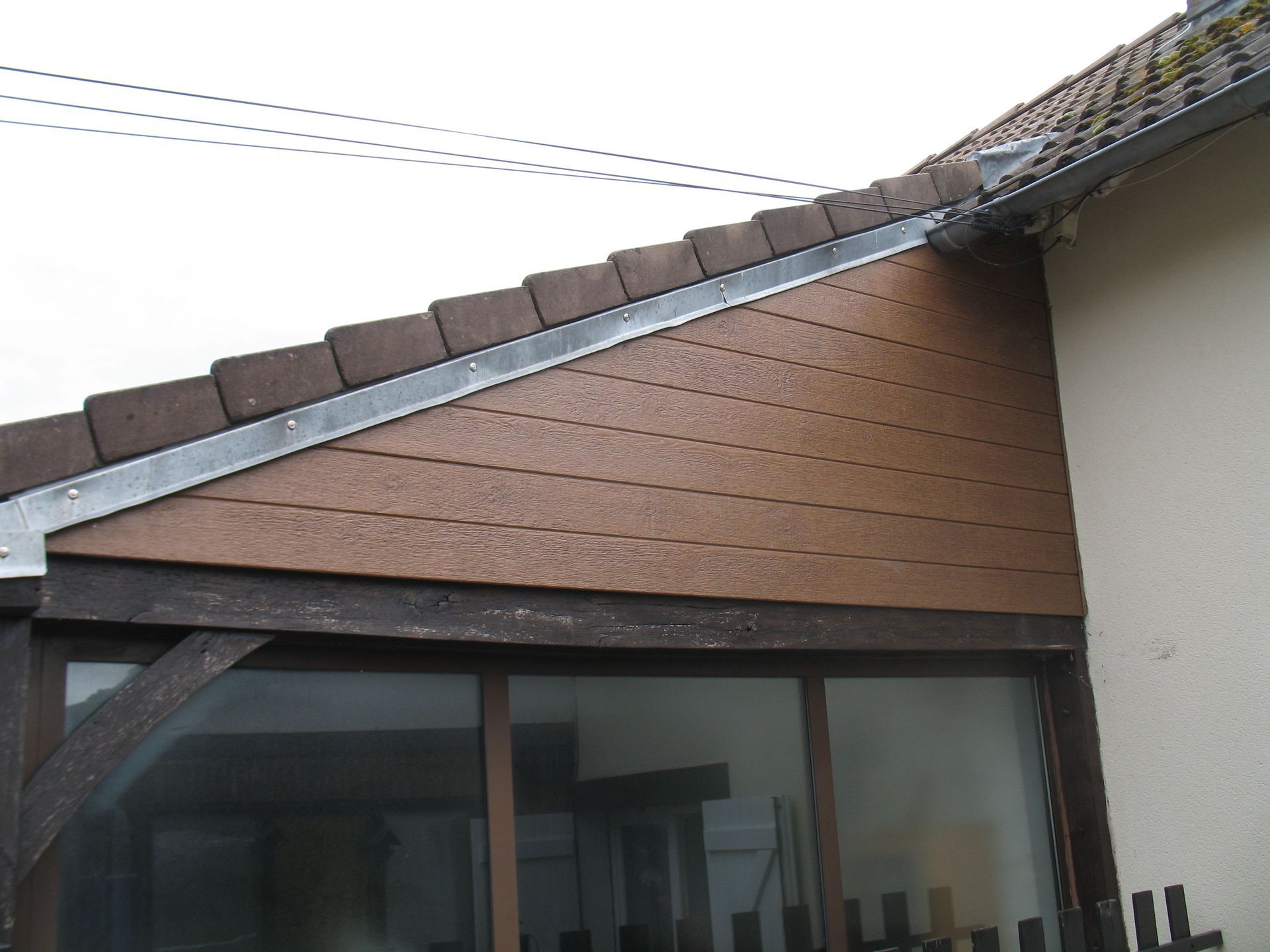 Revêtement en bois brun sur la façade d'un bâtiment, avec une fenêtre en verre, des tuiles brunes et une gouttière en métal.