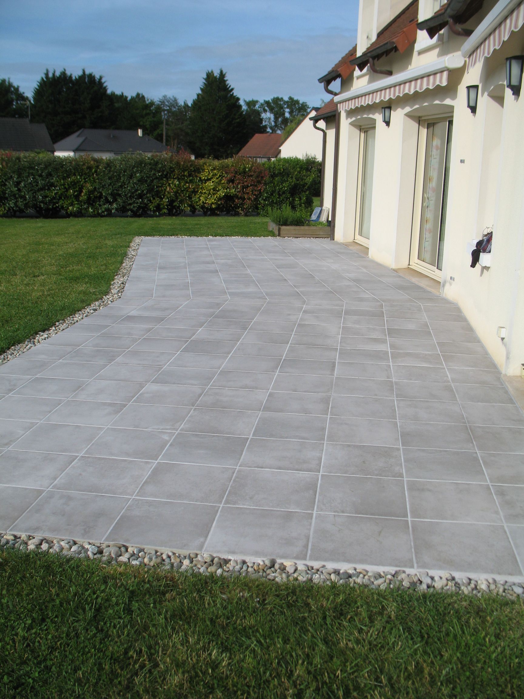 Patio avec carrelage gris à côté d'une maison avec pelouse verte, bordure de gravier.