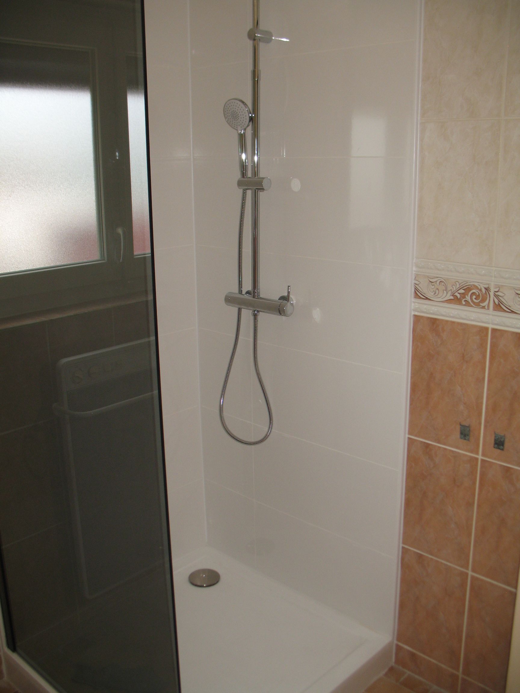 Cabine de douche aux murs blancs, robinetterie chromée et pommeau de douche à main. Mur carrelé marron à droite.