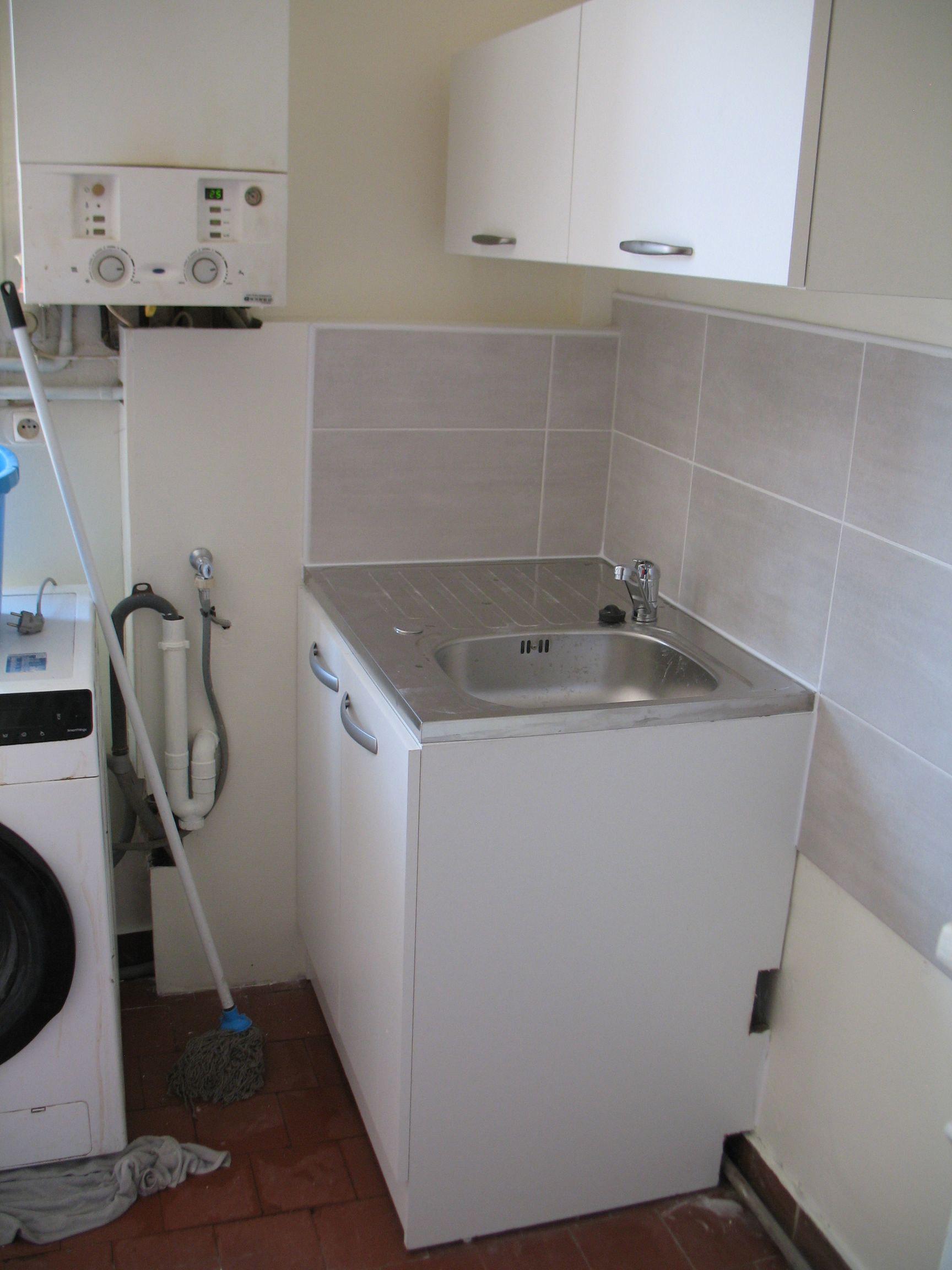 Buanderie avec évier blanc, placards et machine à laver. Balai et tuyau d'arrosage fixés au mur.