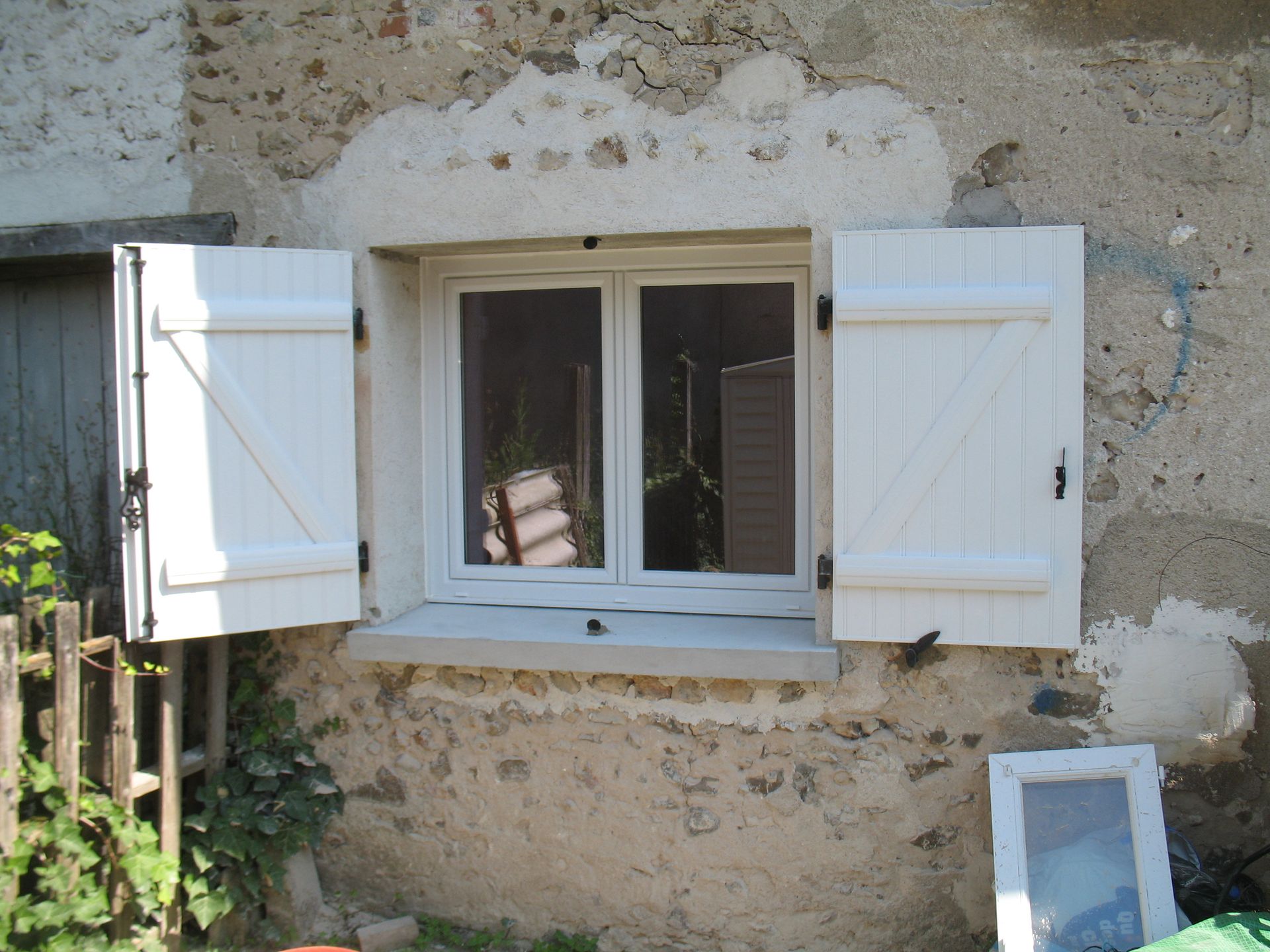 Fenêtre à cadre blanc avec volets ouverts sur la façade d'une maison en pierre. La lumière du soleil se reflète sur la vitre.