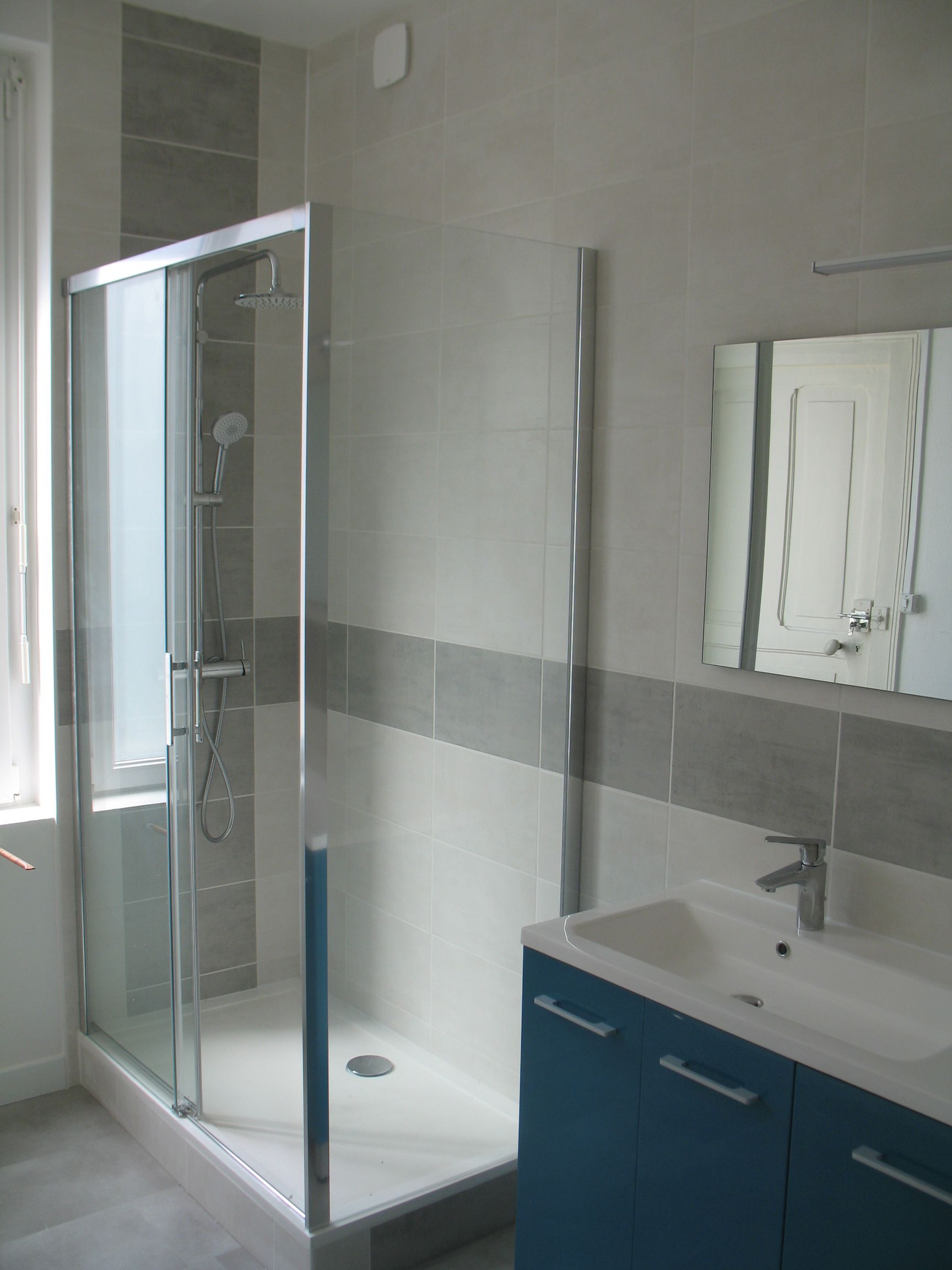 Salle de bain avec douche, meuble-lavabo et miroir. Carrelage gris et bleu, meuble-lavabo bleu, cadre de douche argenté.