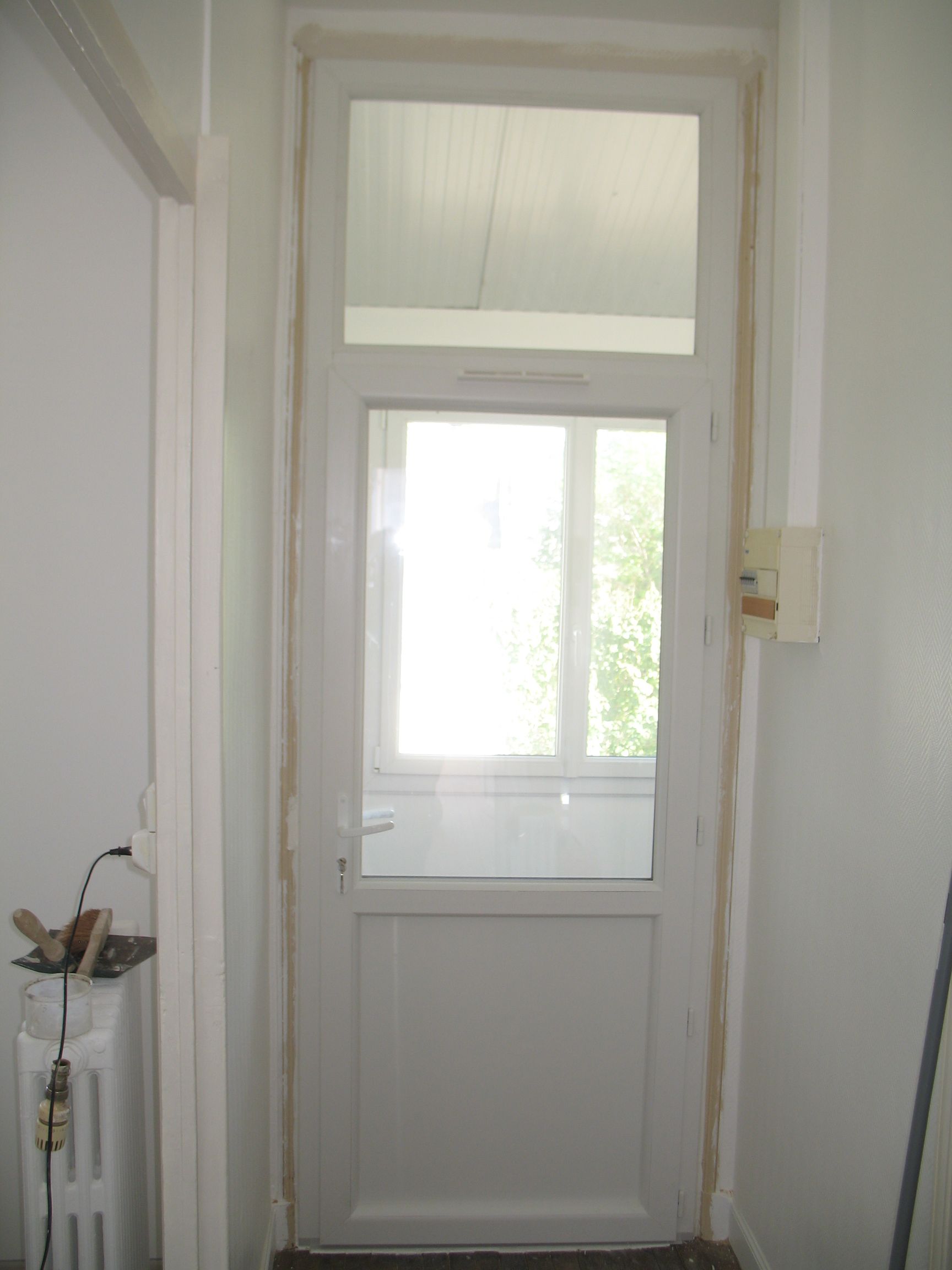 Porte blanche avec fenêtre rectangulaire au-dessus et en dessous, dans un couloir blanc.