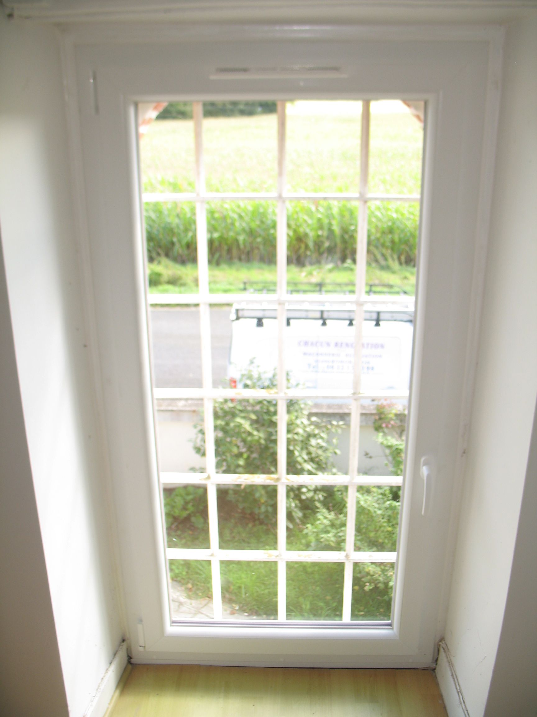 Fenêtre à cadre blanc avec carreaux grillagés, donnant sur la verdure, une route et un panneau.