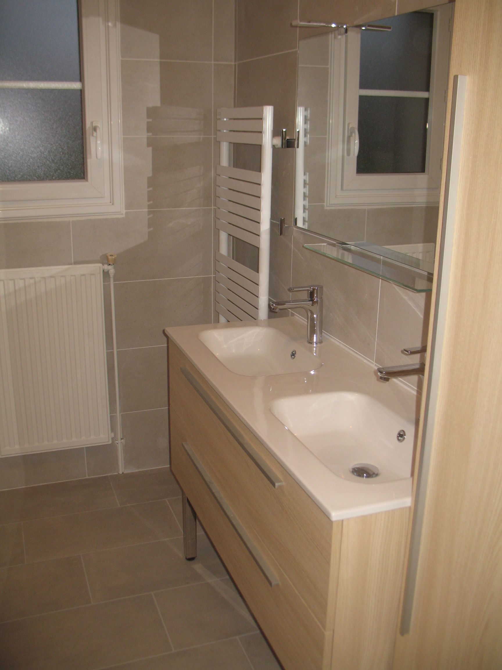 Salle de bains avec meuble à double vasque, sèche-serviettes, miroir et carrelage de couleur claire.