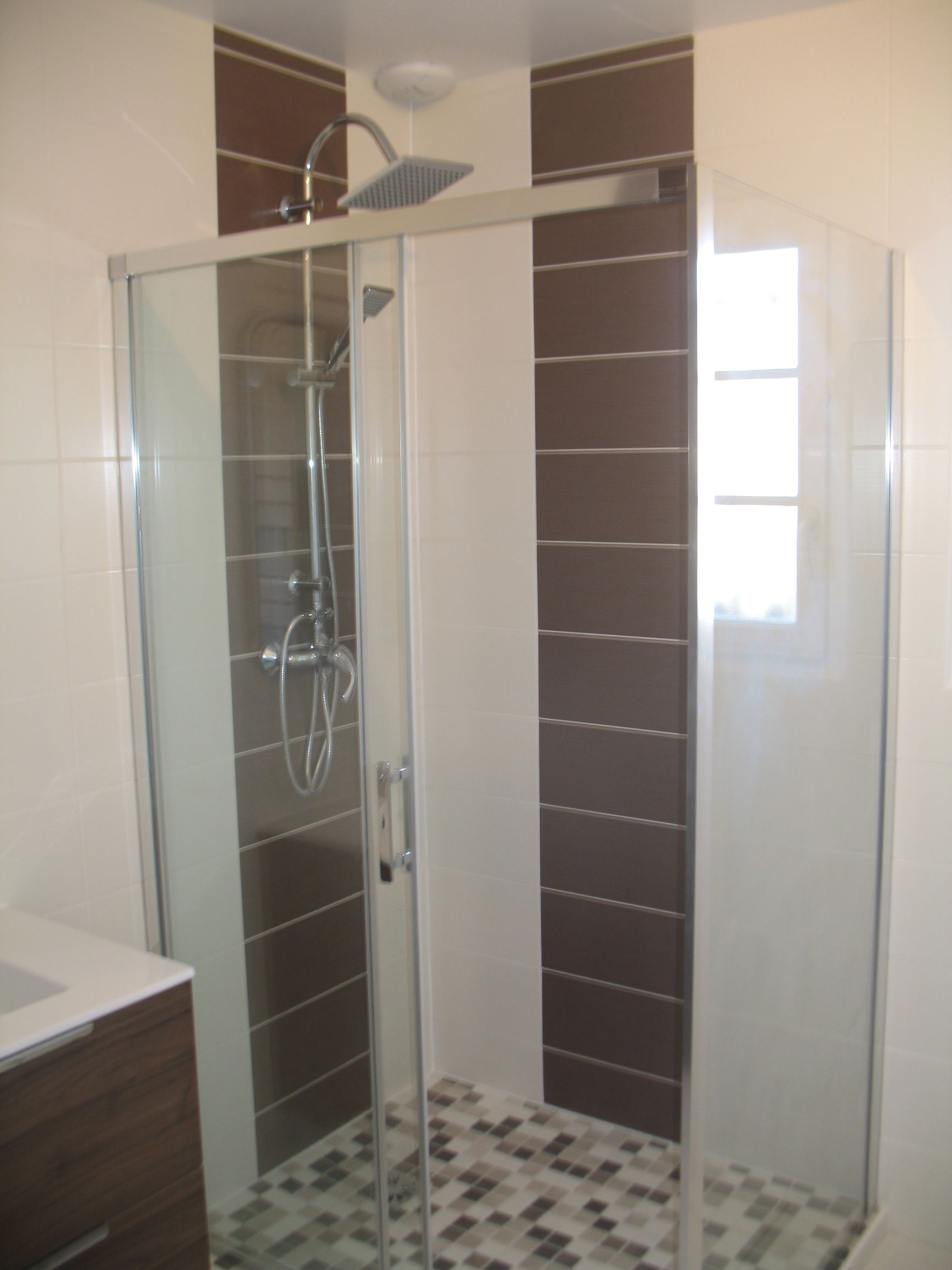 Douche avec porte vitrée, touches de carrelage brun, sol en galets et luminaires chromés.