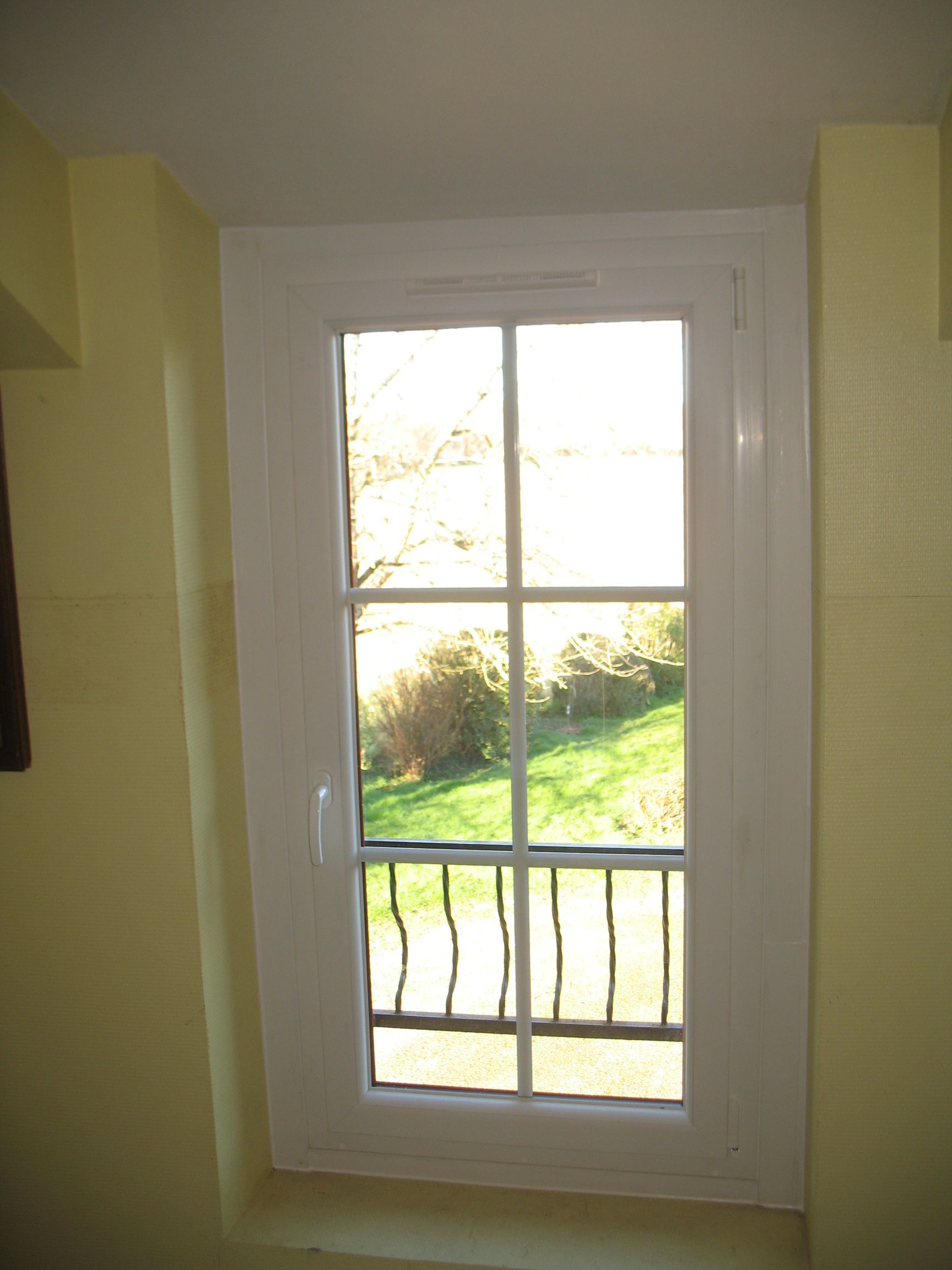 Fenêtre blanche à six carreaux donnant sur une cour herbeuse et un balcon en fer forgé.