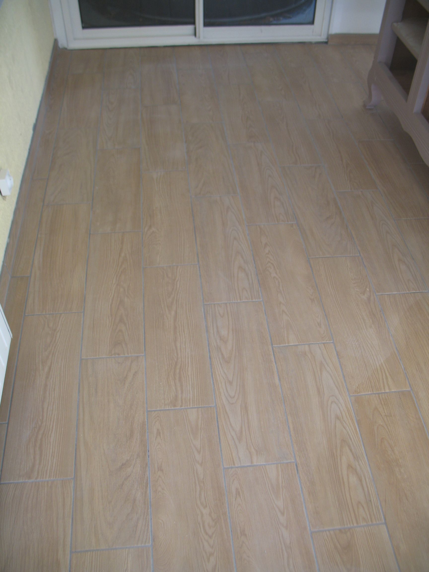 Carrelage aspect bois dans une pièce, menant vers une porte vitrée ; une armoire beige se trouve à droite.