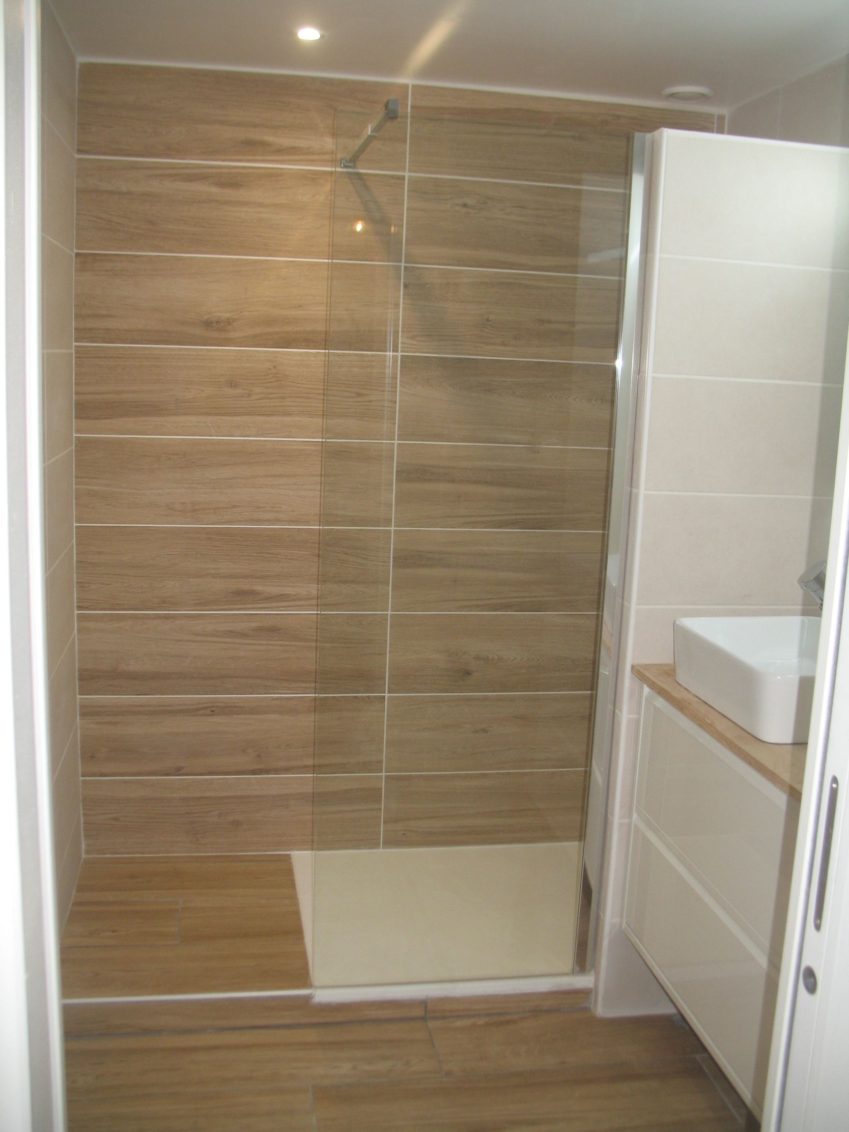Salle de bain avec murs et sol en carrelage imitation bois, cabine de douche en verre, vanité blanche et luminaires modernes.