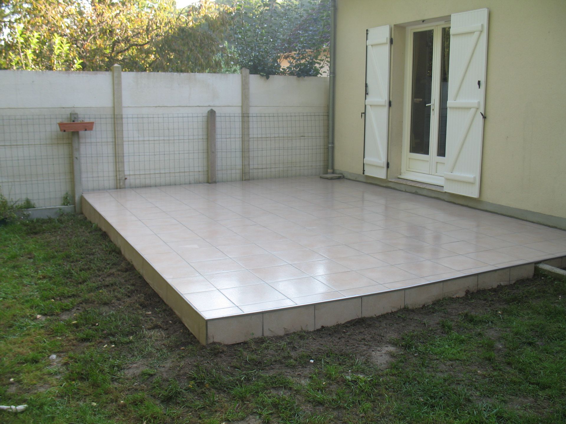 Patio carrelé à l'extérieur d'une maison avec portes-fenêtres ouvertes et volets blancs ; un mur en béton en arrière-plan.