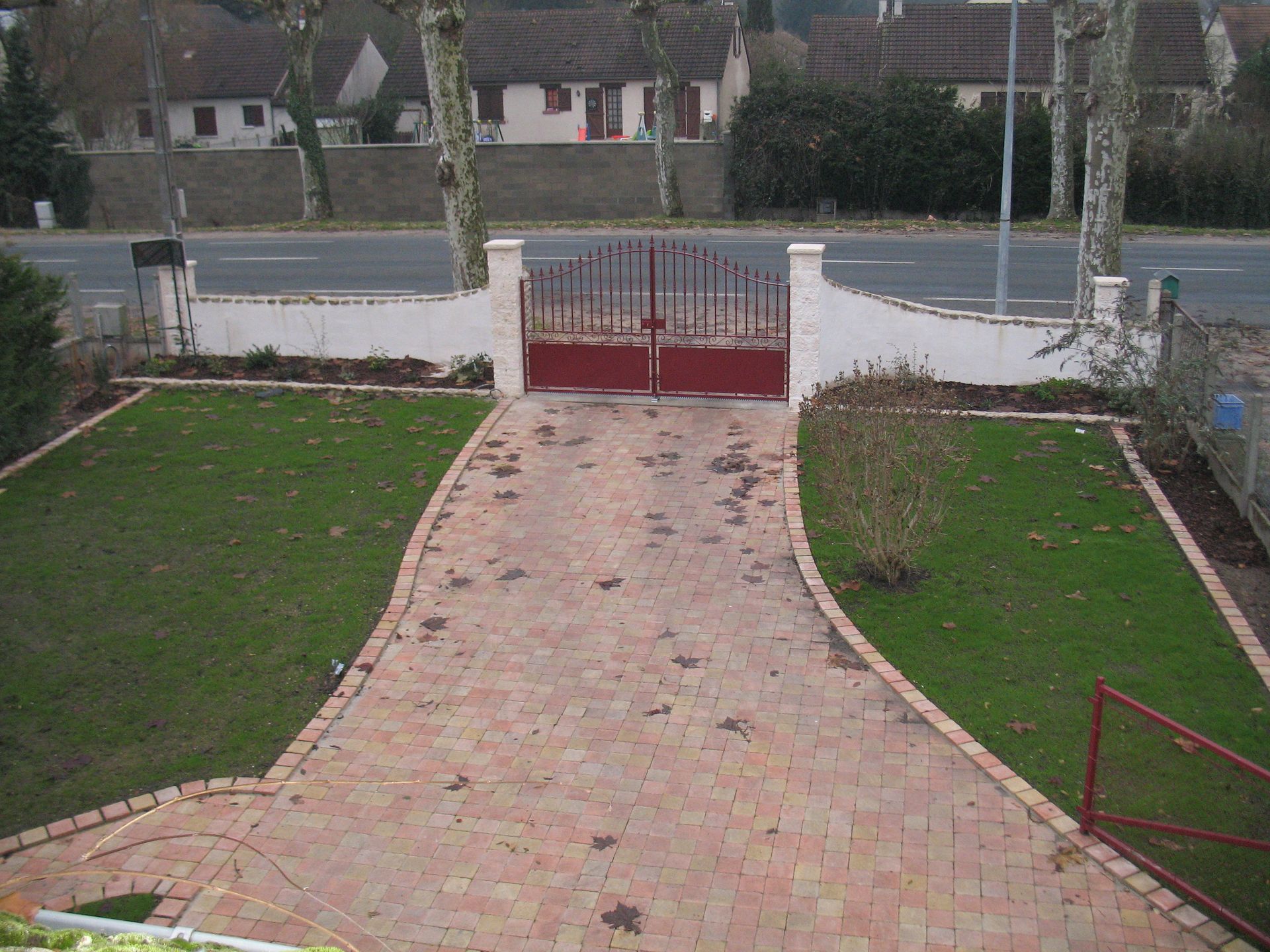 Allée en briques rouges menant au portail et au mur bordeaux, flanquée d'herbe et de route.