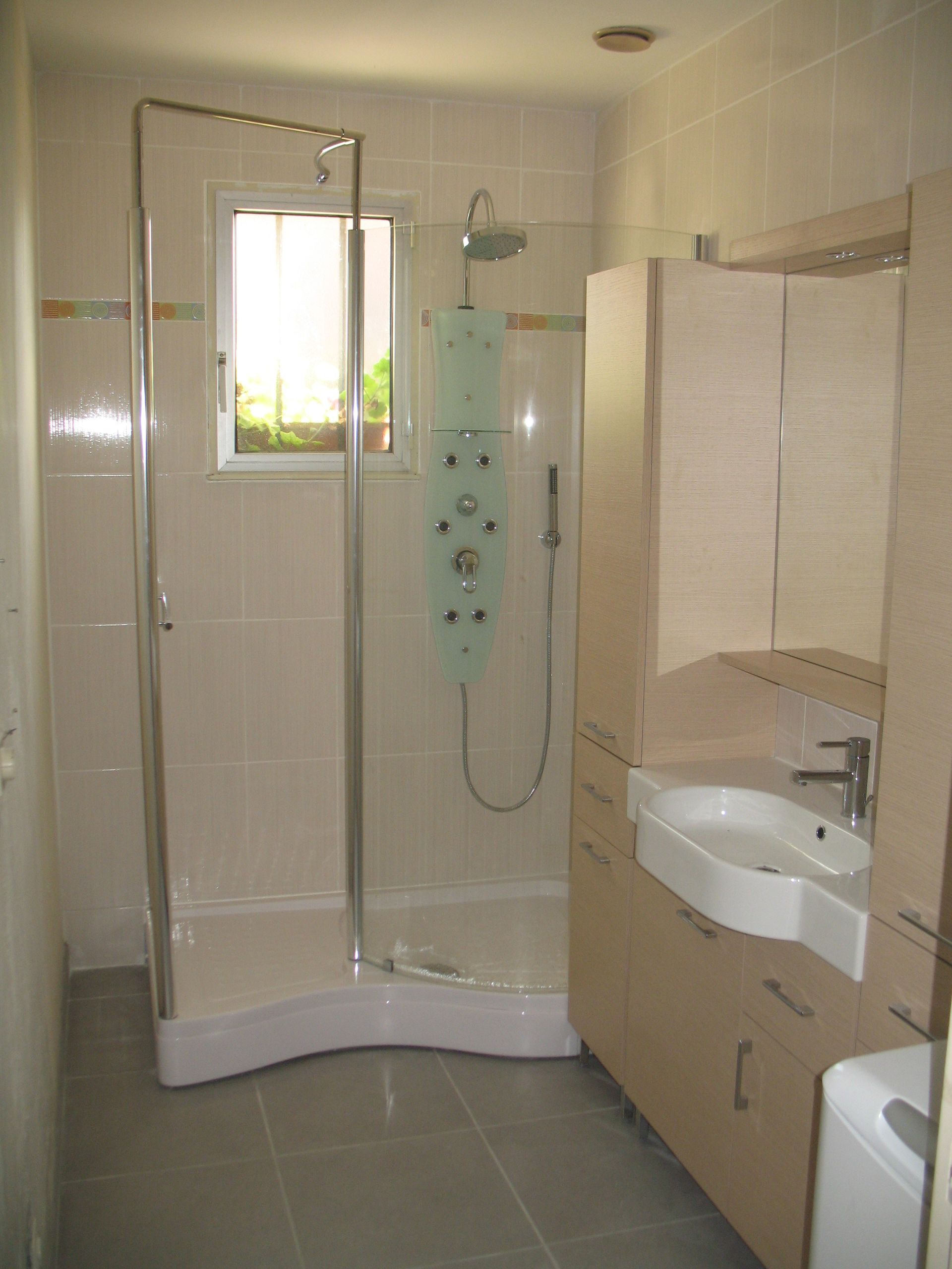 Salle de bain avec douche, meuble-lavabo et meubles beige clair. Petite fenêtre à gauche.