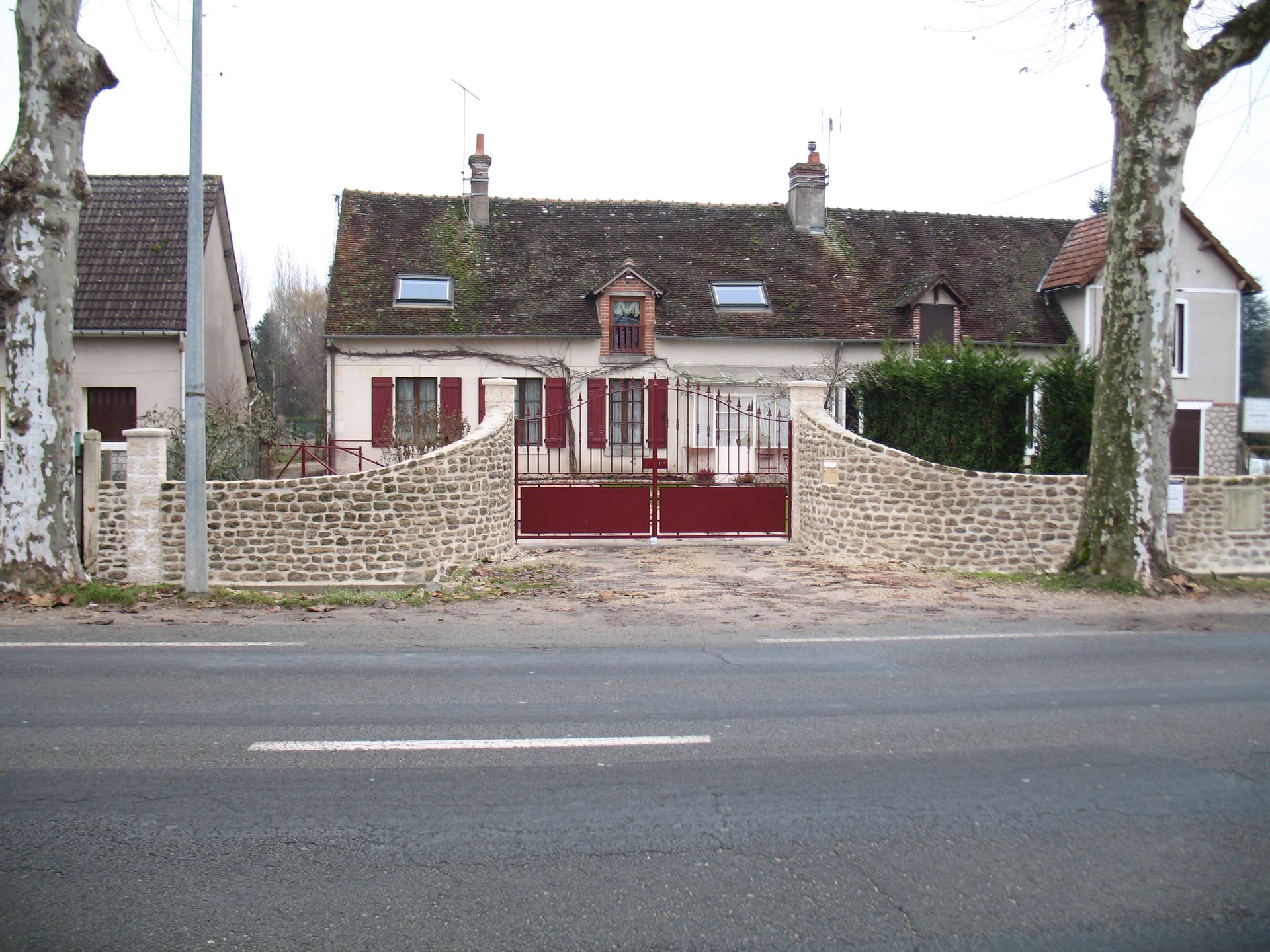 Une maison avec un portail rouge et un mur de briques à côté d'une route, des arbres sur les côtés, un ciel couvert.