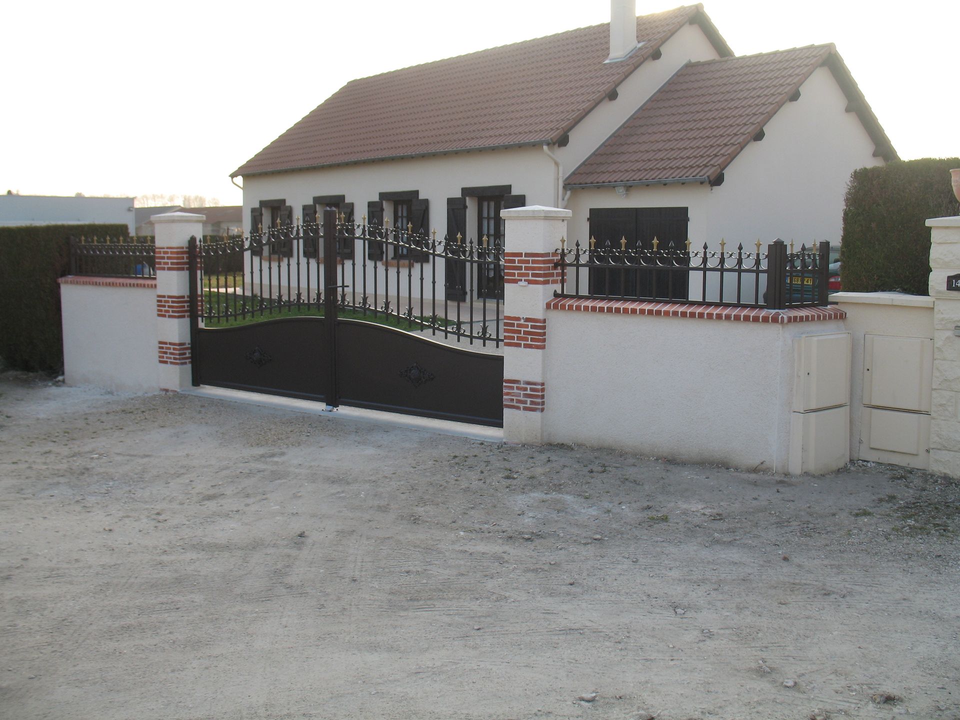 Une maison avec une clôture en briques et blanche, un portail noir et une allée en gravier.