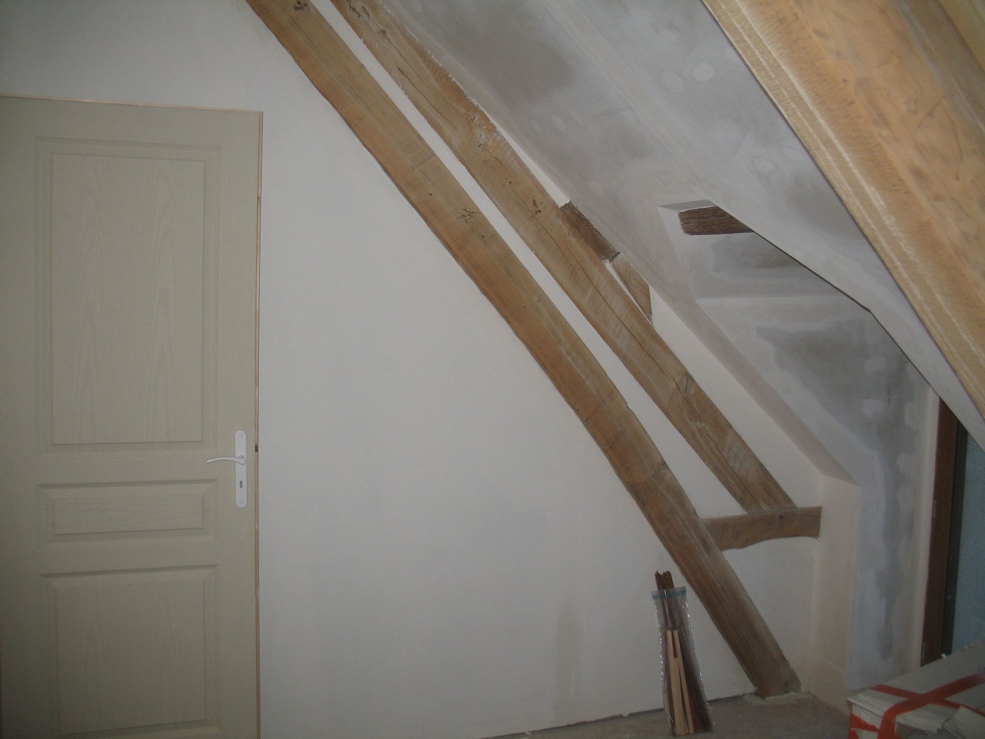Intérieur d'une pièce avec plafond incliné et poutres en bois. Une porte se trouve à gauche. Murs blancs.