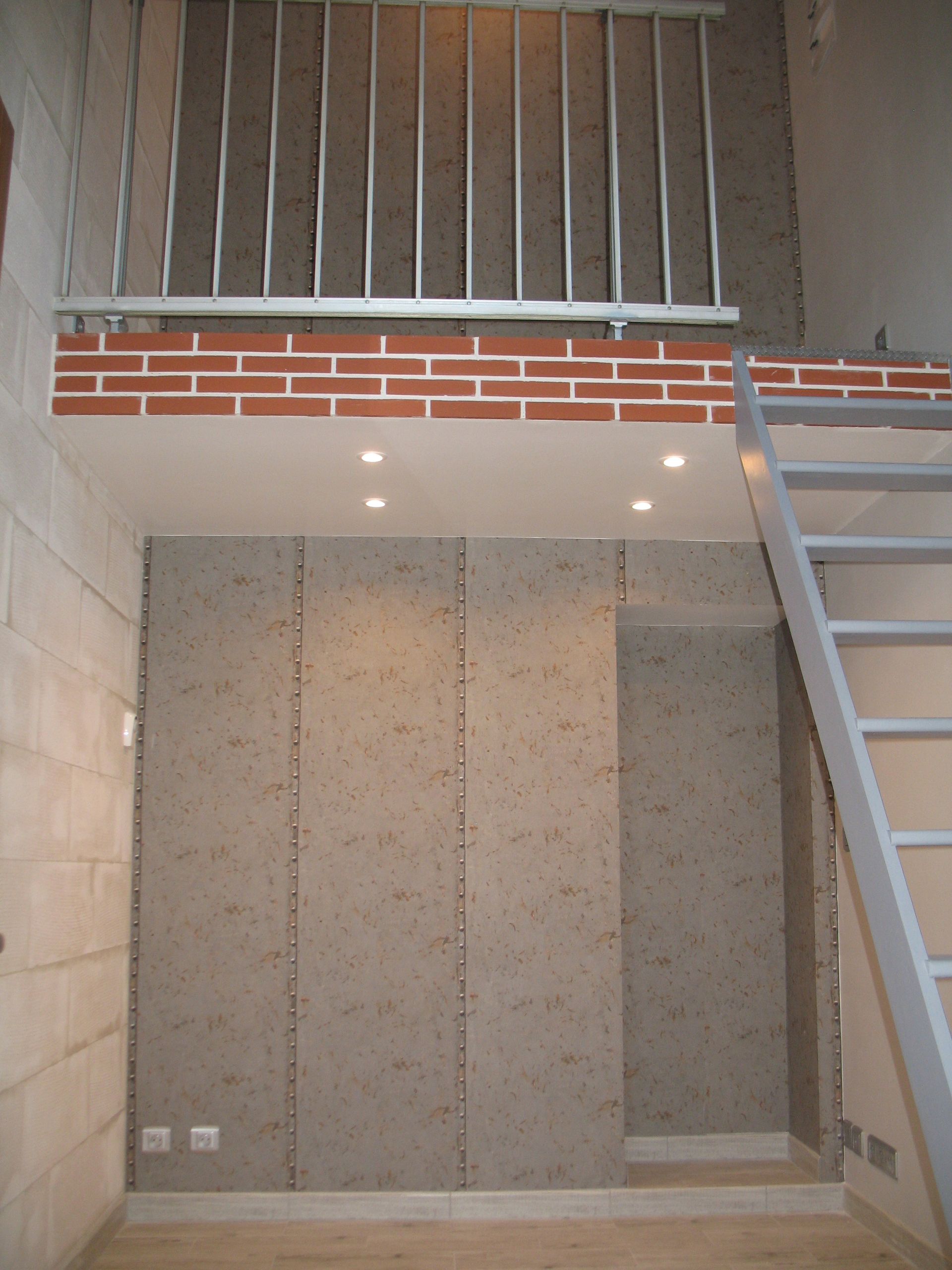 Loft avec mur en briques, rampe et échelle en métal. Boiseries grises et petits spots encastrés.