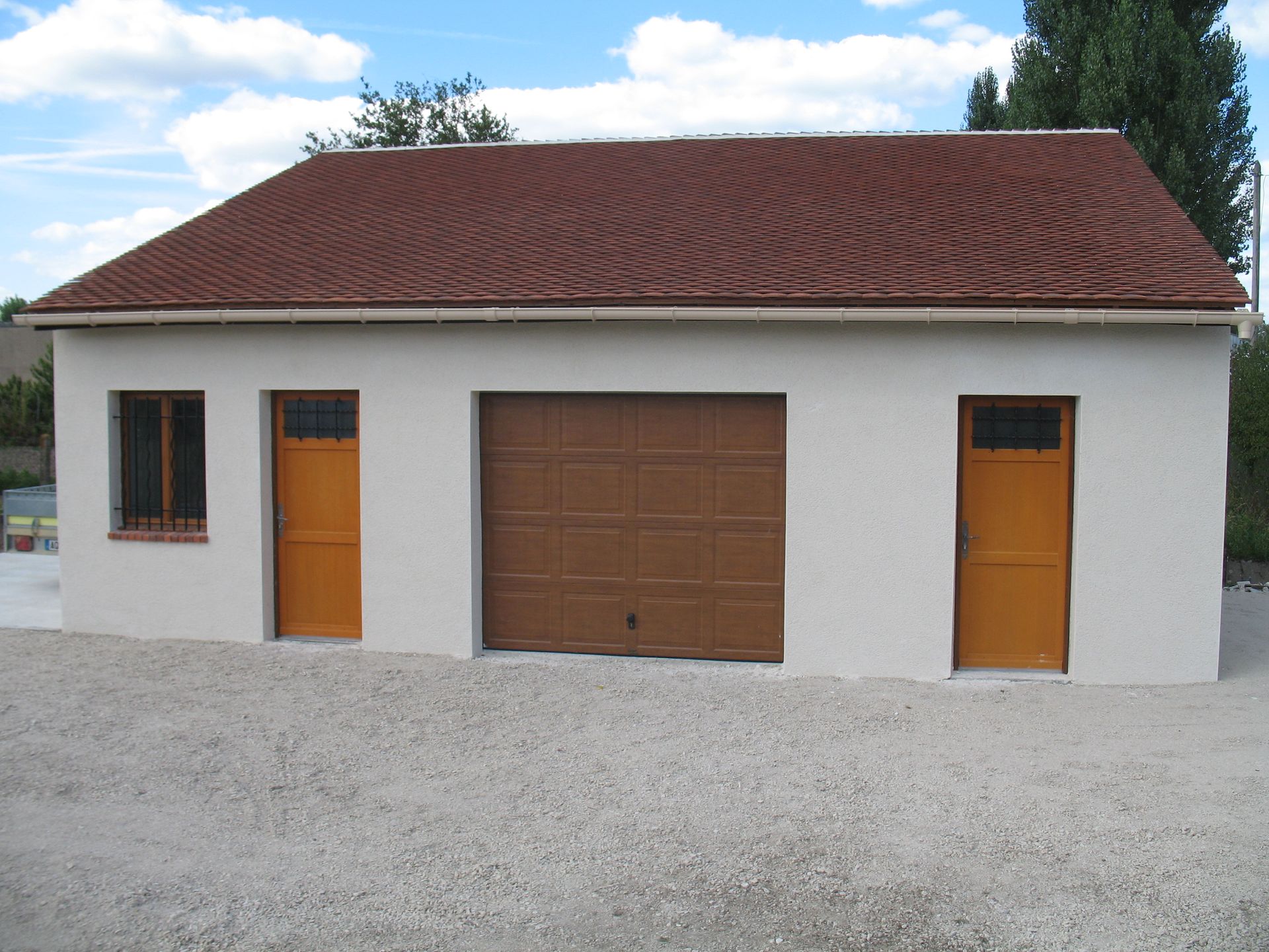 Garage blanc avec porte marron, portes orange et toit en tuiles rouges sur gravier.