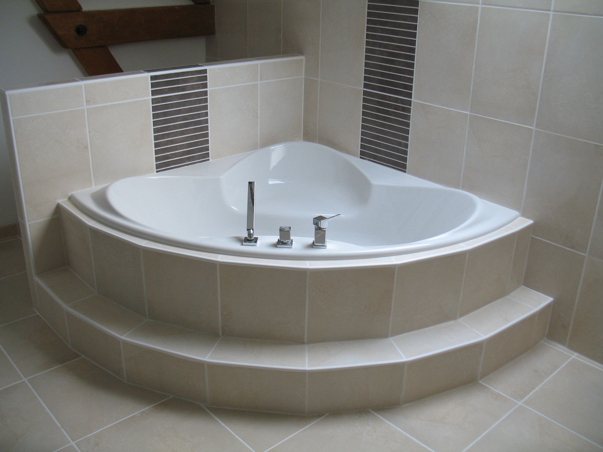 Baignoire jacuzzi d'angle dans une alcôve carrelée avec deux marches, carrelage beige clair et rayures décoratives.