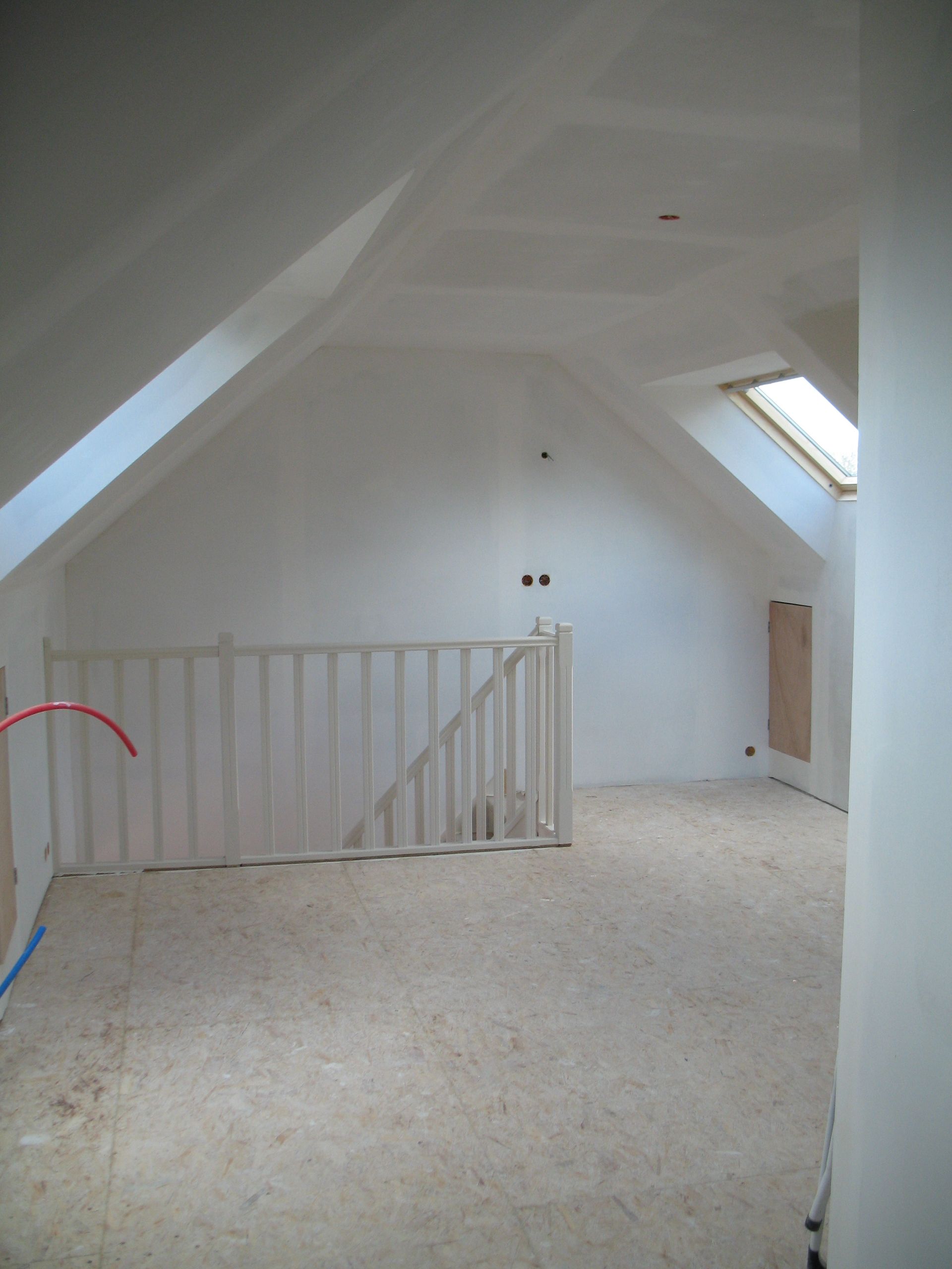Intérieur d'une chambre mansardée inachevée avec plafonds en pente, sol non fini et une petite balustrade partielle.