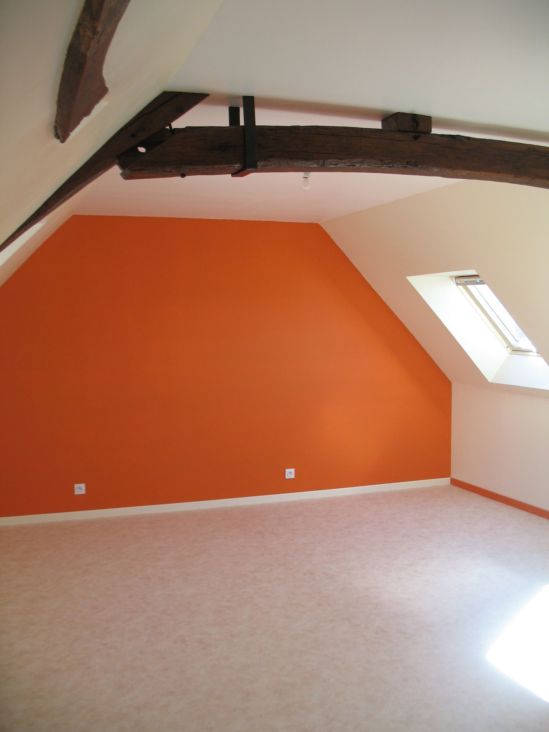Mur orange dans une pièce mansardée avec plafonds inclinés, puits de lumière et poutres en bois apparentes.