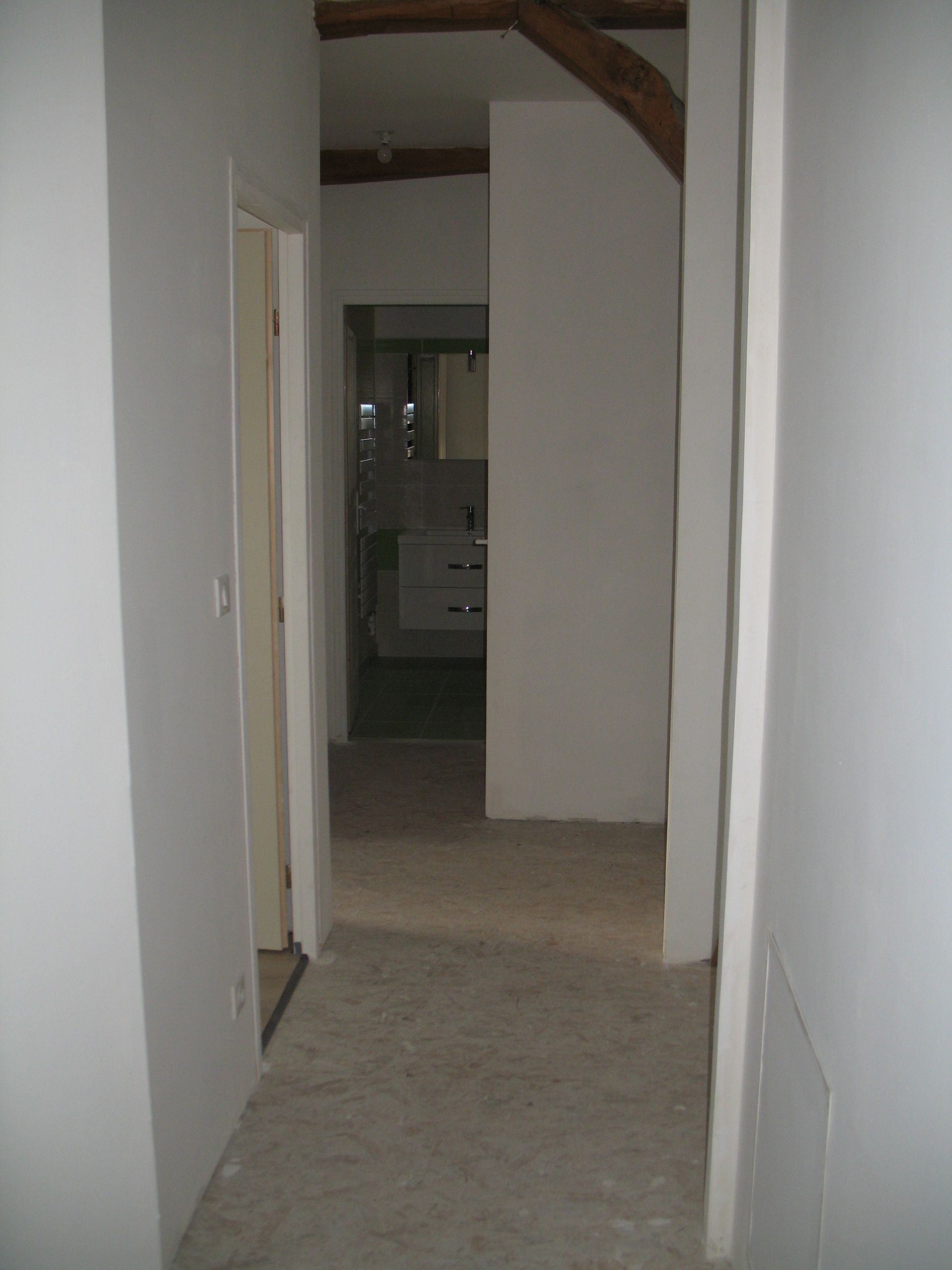 Couloir étroit avec murs et portes blancs, menant à une salle de bain avec armoires blanches.