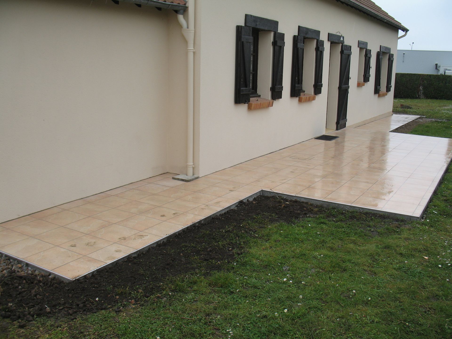 Terrasse carrelée beige à côté d'une maison beige aux volets noirs, fraîchement posée.
