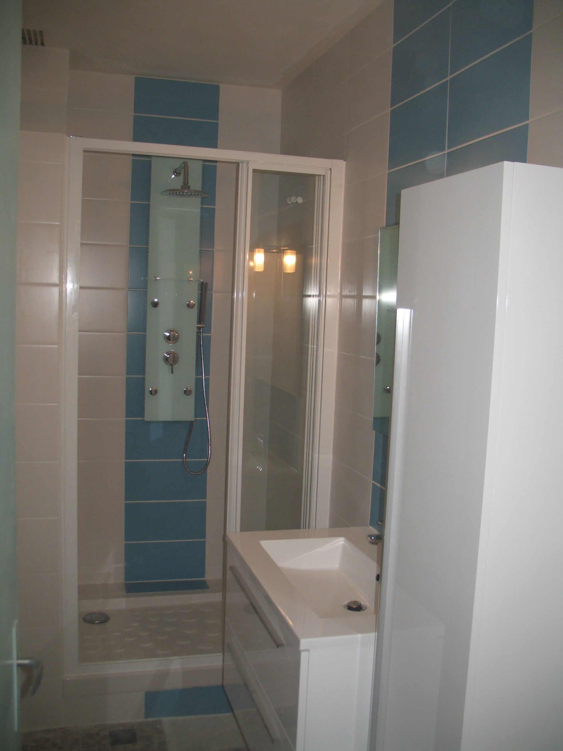 Salle de bain avec carrelage blanc et bleu, porte de douche en verre, lavabo et armoire blanche.