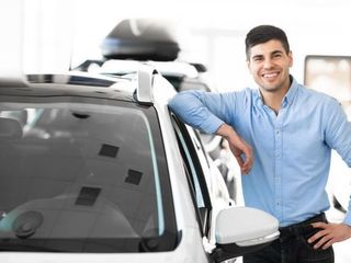 Un hombre con una camisa azul está apoyado en un coche blanco.