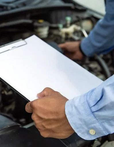 Un hombre mira debajo del capó de un automóvil mientras sostiene un portapapeles.