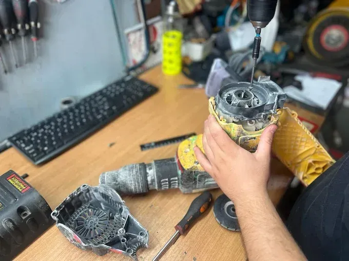 Una persona utiliza un destornillador eléctrico para reparar un taladro percutor amarillo en un banco de trabajo desordenado.